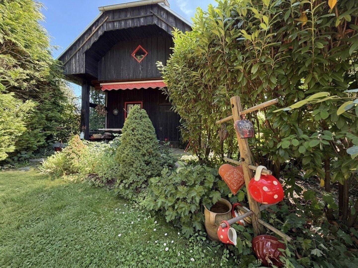 Rustikales Holzhaus mit roter Markise, umgeben von einem blühenden Garten mit Dekorationen.