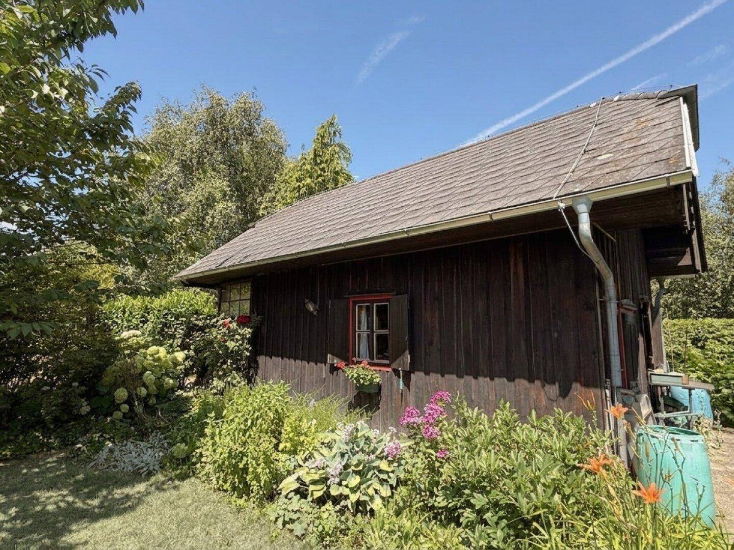 Seitenansicht des Holzhauses mit blühenden Pflanzen und einem kleinen Fenster mit roten Fensterläden.