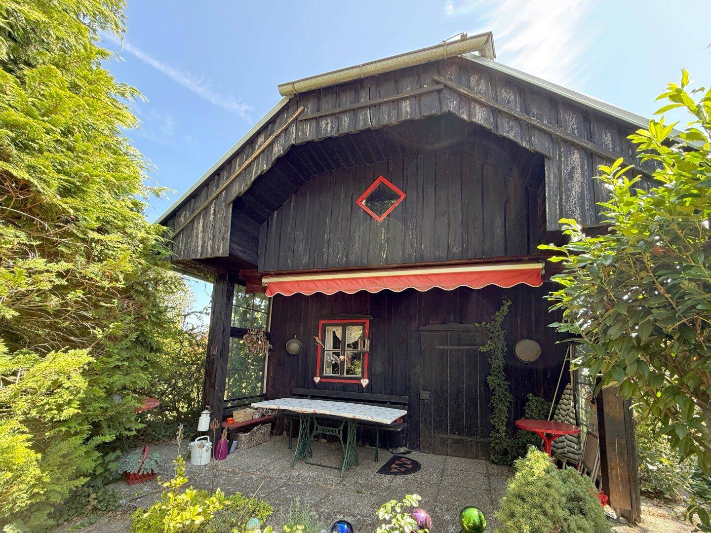 Sonnige Terrasse mit rustikalem Tisch und Markise vor der dunklen Holzfassade des Hauses.