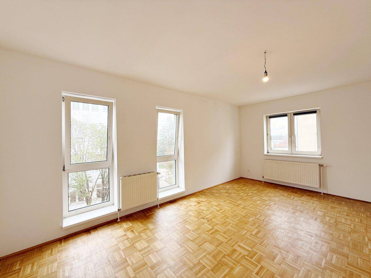 Heller, leerer Raum mit Fischgrätparkett und großen Fenstern.