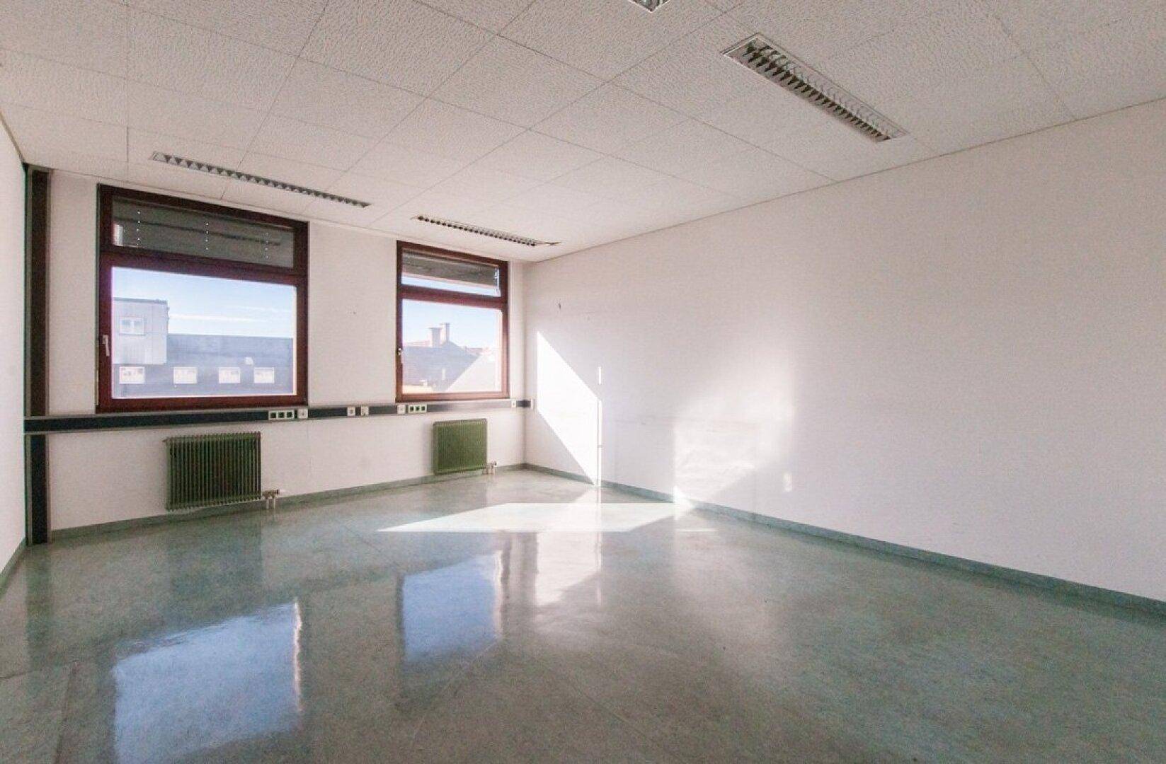 Leeres Büro mit hellen Wänden, zwei Fenstern und einem glänzenden grauen Boden, der das Sonnenlicht reflektiert.