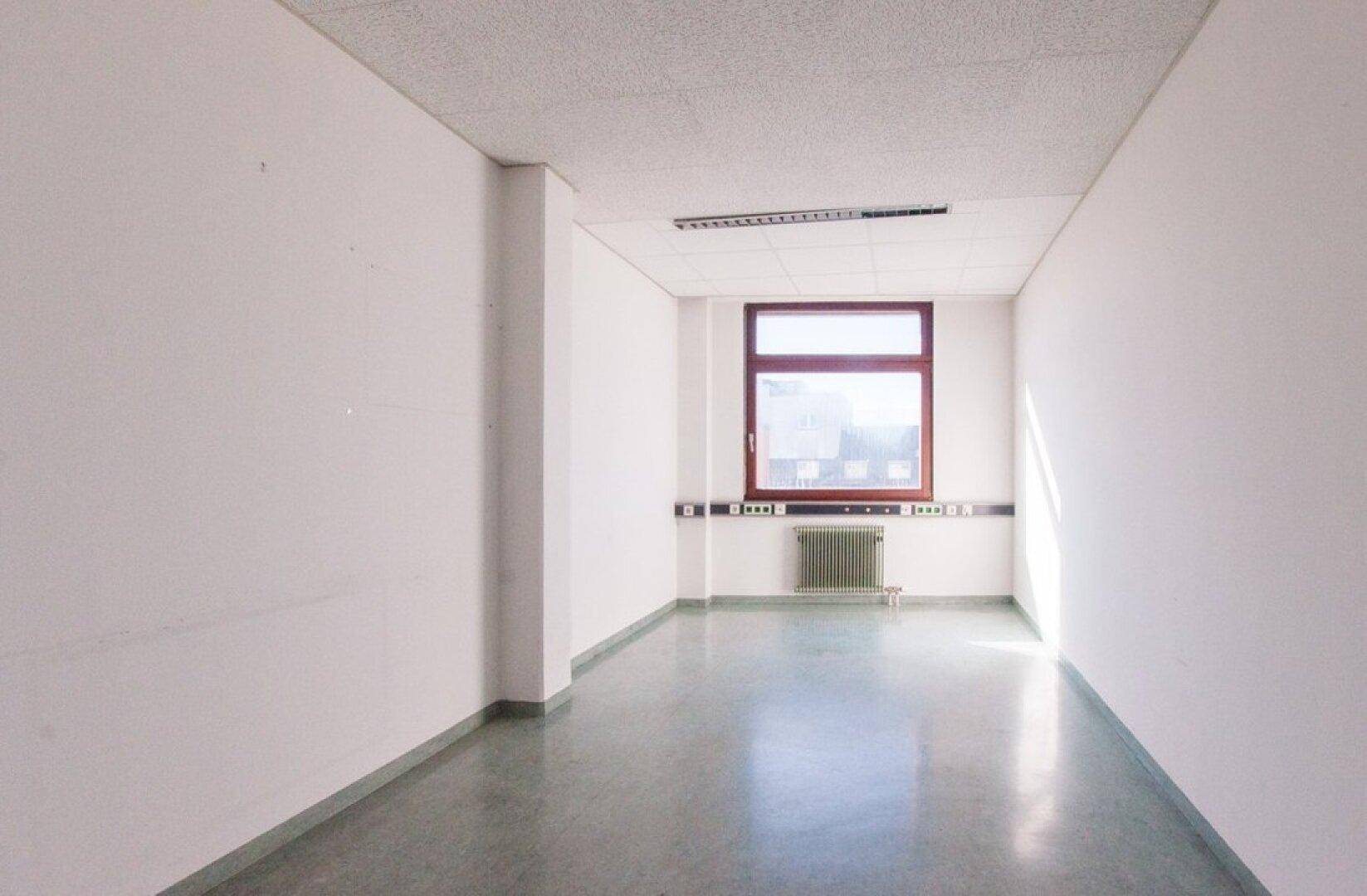 Schmaler Büroraum mit einem Fenster, weißen Wänden und einem glänzenden grauen Boden, der das Licht reflektiert.