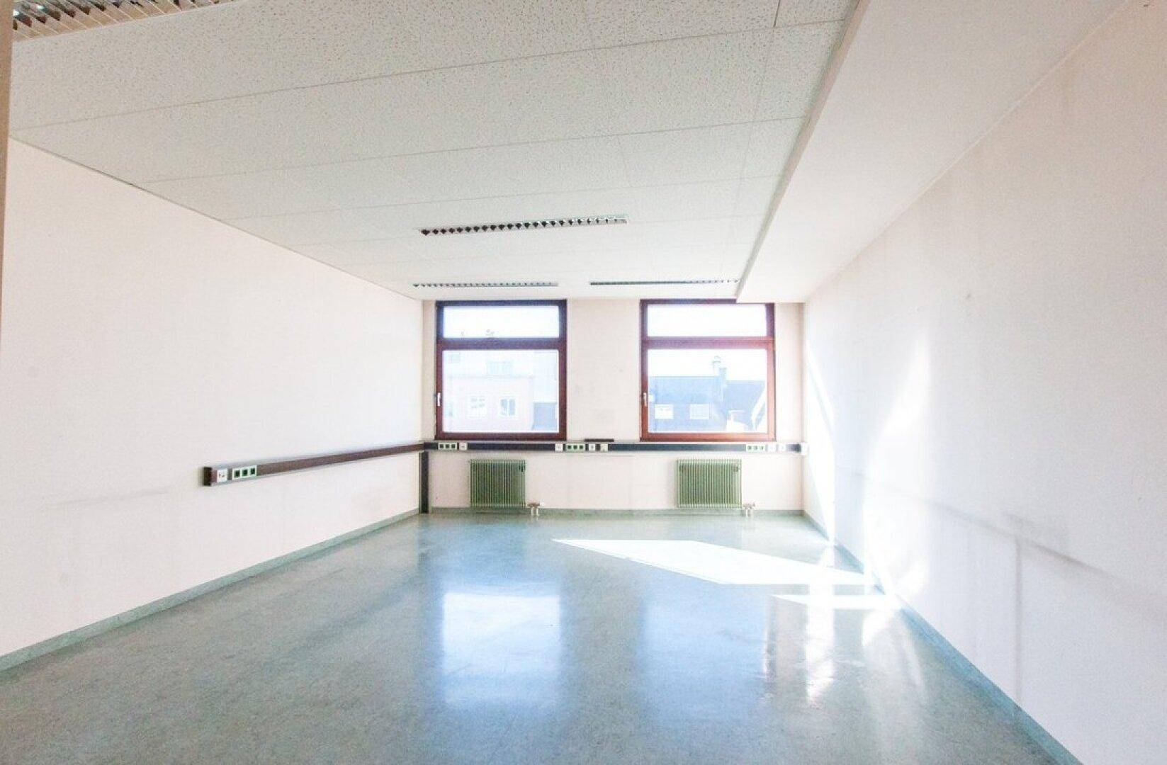 Helles Büro mit zwei Fenstern, weißen Wänden und einem glänzenden grauen Boden, der das Sonnenlicht reflektiert.
