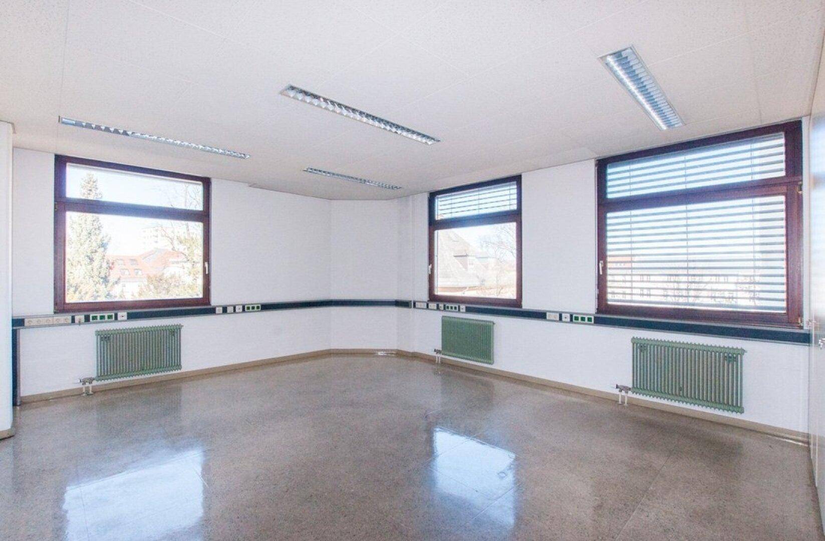 Geräumiges Büro mit drei Fenstern, weißen Wänden und einem glänzenden grauen Boden, der das Sonnenlicht reflektiert.