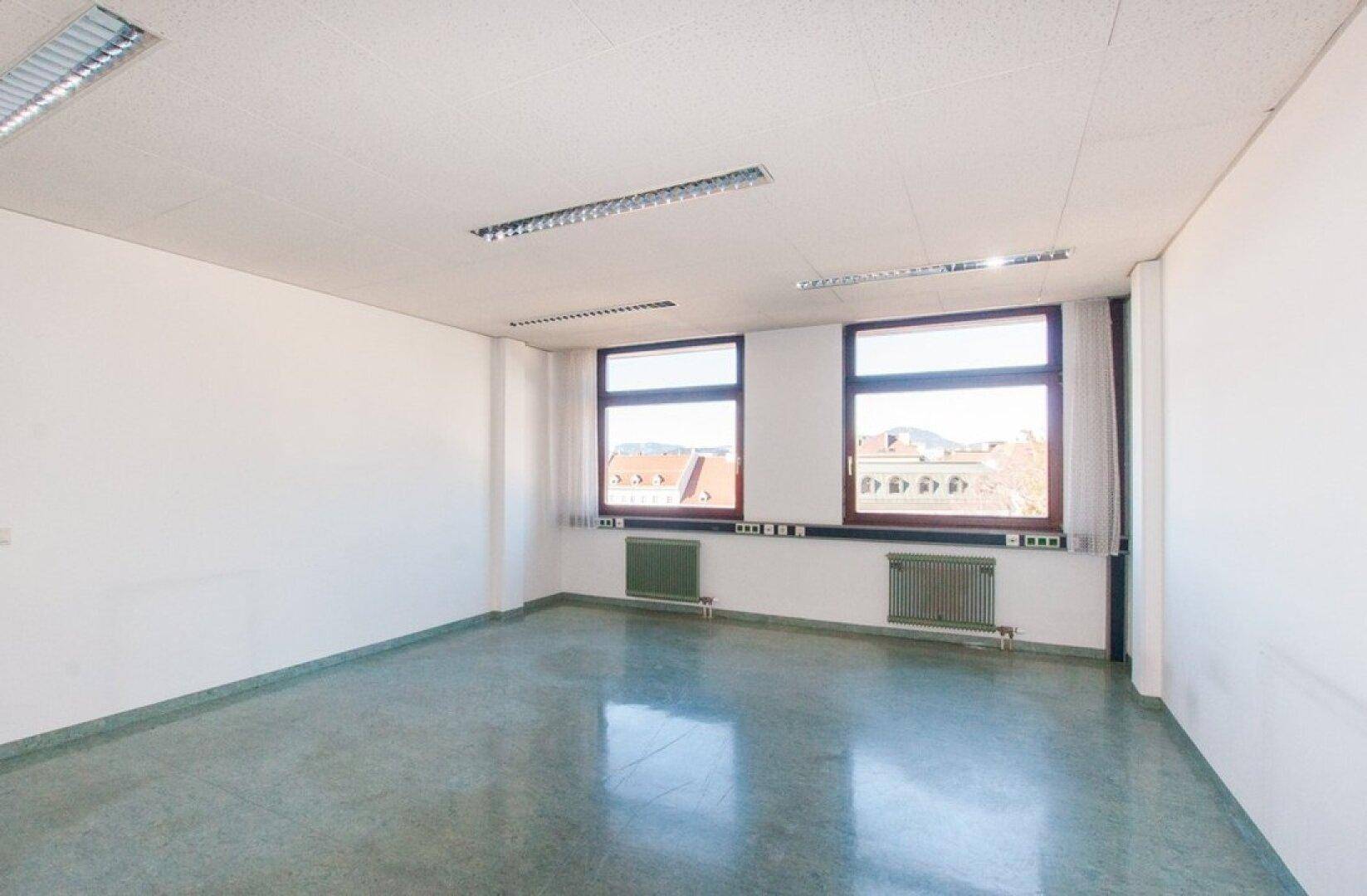 Geräumiges Büro mit zwei Fenstern, weißen Wänden und einem glänzenden grauen Boden, der das Sonnenlicht reflektiert.