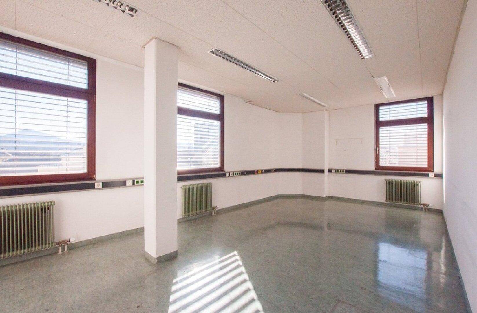 Geräumiges Büro mit drei Fenstern, weißen Wänden und einem glänzenden grauen Boden, der das Sonnenlicht reflektiert.