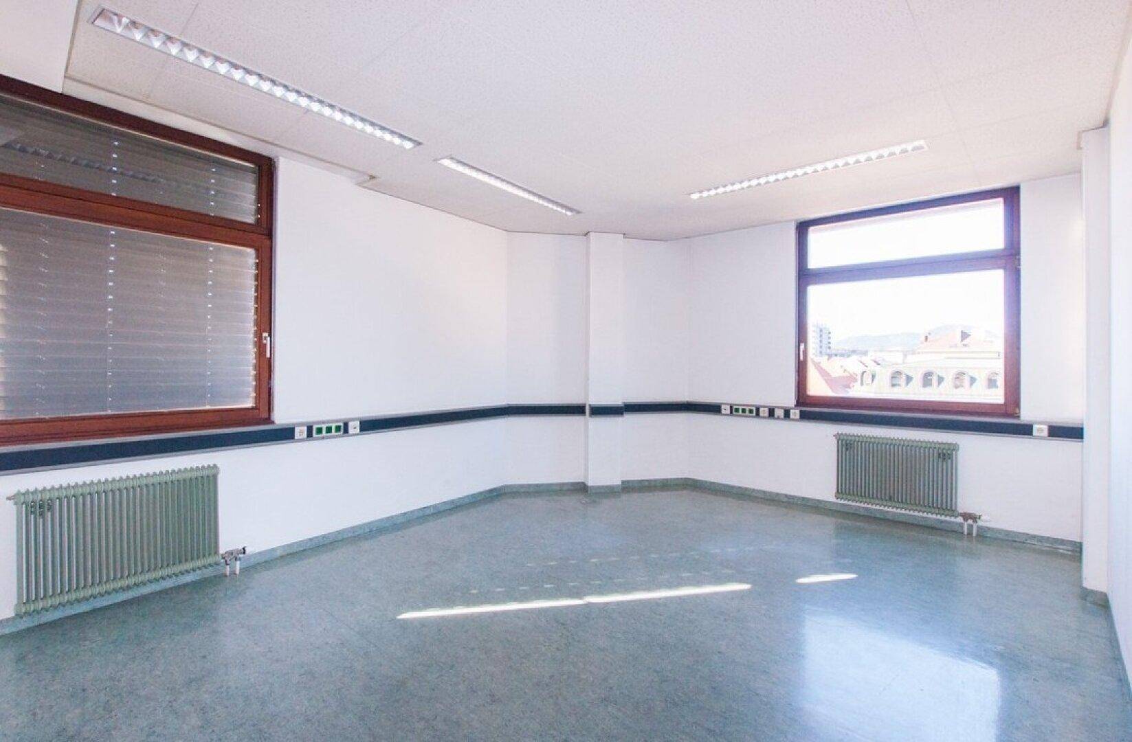 Helles Büro mit zwei Fenstern, weißen Wänden und einem glänzenden grauen Boden, der das Sonnenlicht reflektiert.