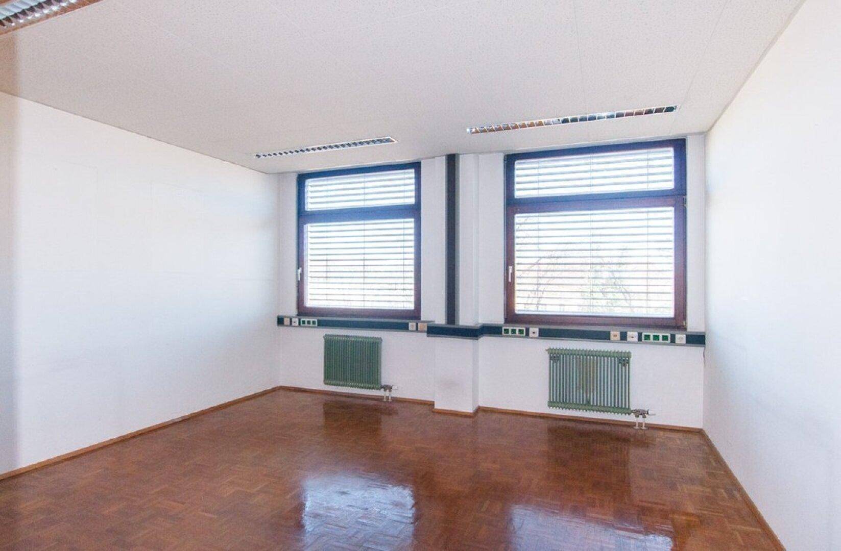 Helles Büro mit zwei Fenstern, weißen Wänden und einem warmen Parkettboden, der das Sonnenlicht reflektiert.