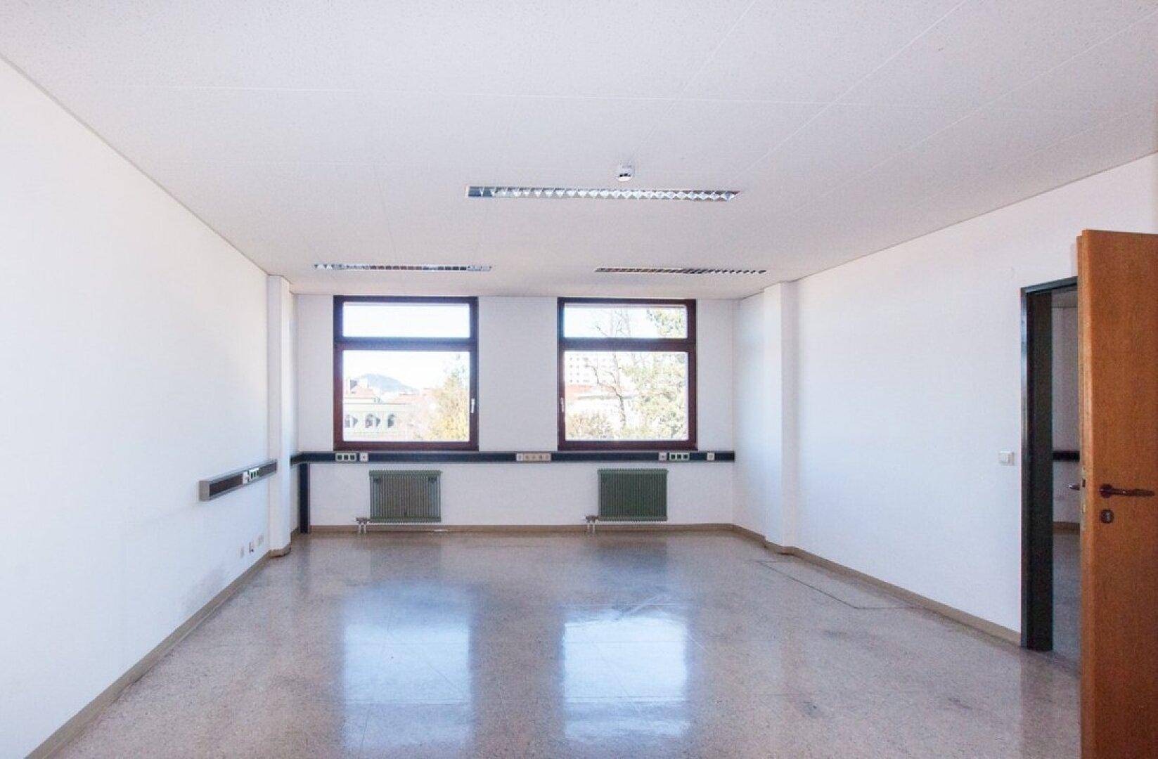 Leeres Büro mit zwei Fenstern, weißen Wänden und einem glänzenden grauen Boden, der das Sonnenlicht reflektiert.