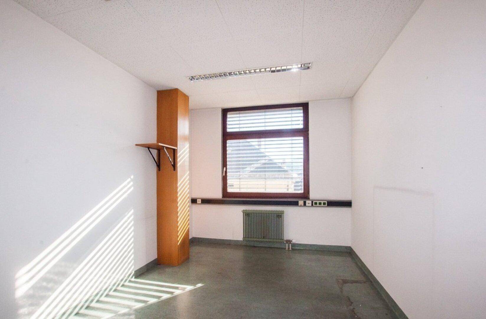 Schmaler Büroraum mit einem Fenster, weißen Wänden und einem glänzenden grauen Boden, der das Licht reflektiert.