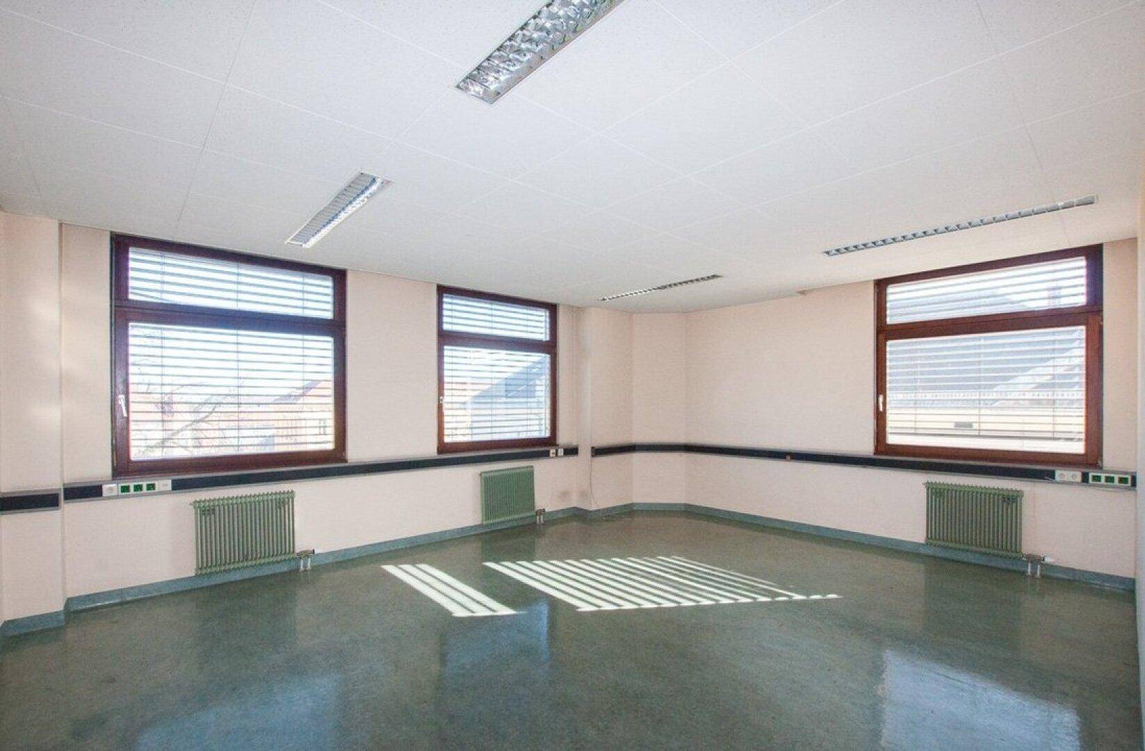 Geräumiges Büro mit drei Fenstern, weißen Wänden und einem glänzenden grauen Boden, der das Sonnenlicht reflektiert.