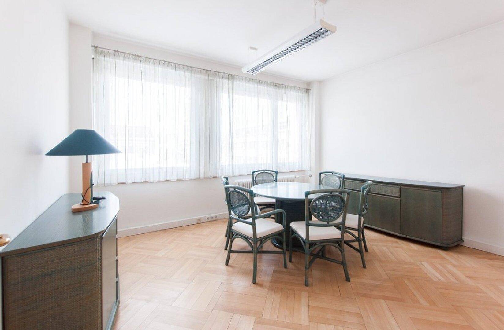 Konferenzraum mit Fischgrätparkett, rundem Tisch und sechs Stühlen, beleuchtet durch große Fenster.