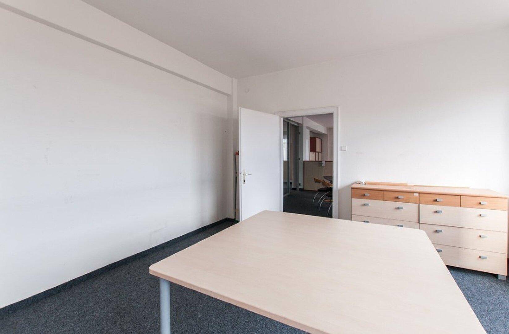 Einfaches Büro mit hellem Tisch, Kommode und einer offenen Tür zu einem weiteren Raum.