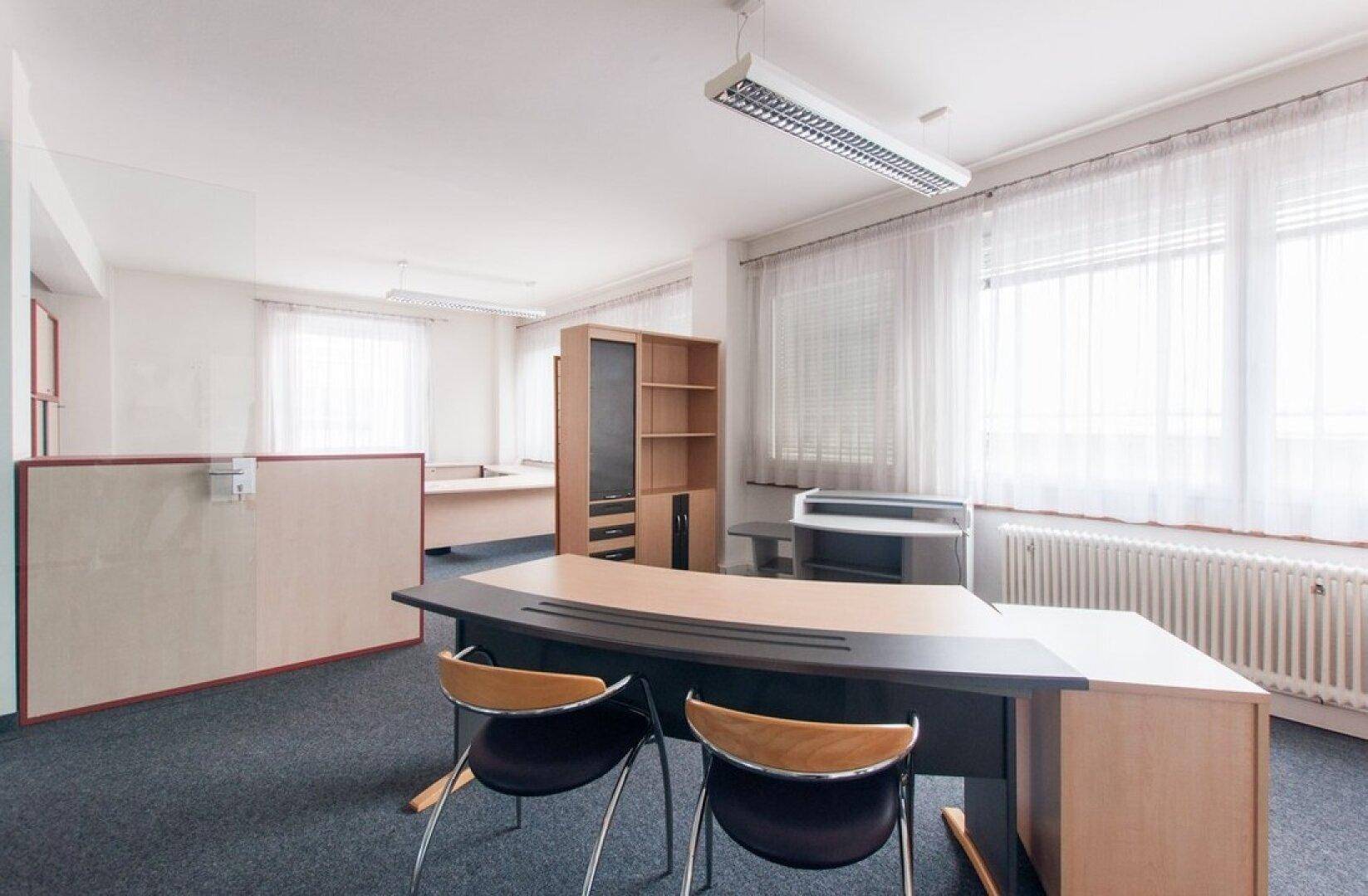 Helles Büro mit großem Schreibtisch, Bürostühlen und Teppichboden, beleuchtet durch große Fenster.