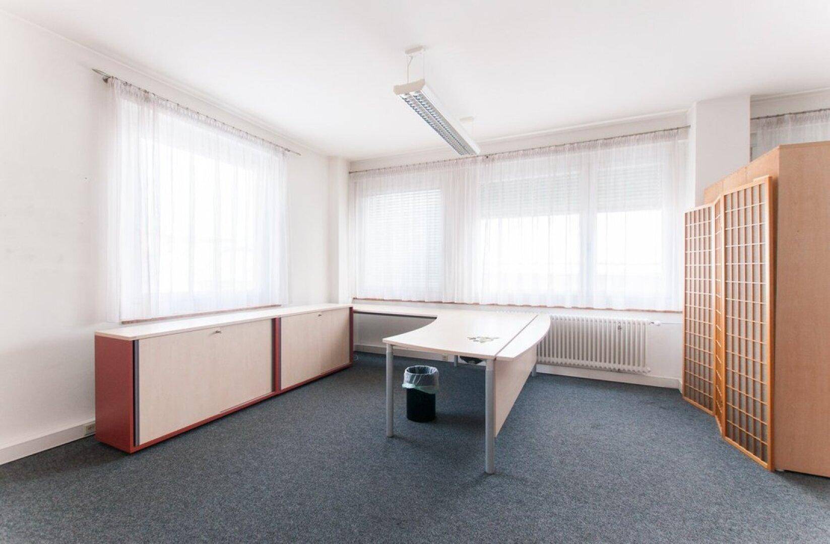 Geräumiges Büro mit Teppichboden, einem geschwungenen Schreibtisch und niedrigen Schränken entlang der Fensterfront.