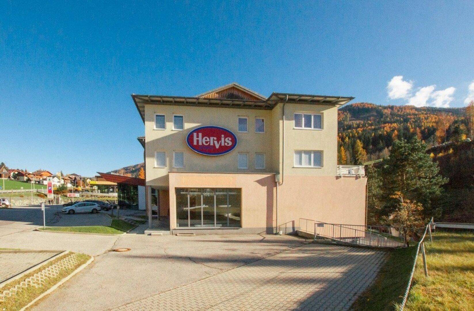 Gebäudeansicht eines Gewerbeobjekts mit Hervis-Logo und Parkplätzen vor einer bergigen Landschaft.