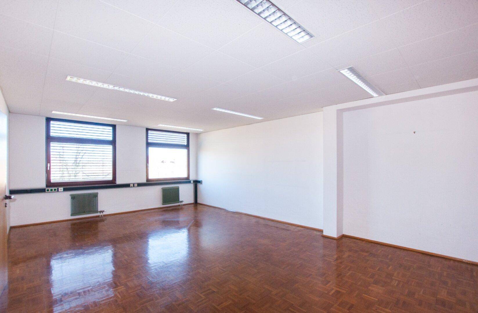 Geräumiges Büro mit zwei Fenstern, weißen Wänden und einem warmen Parkettboden, der das Sonnenlicht reflektiert.