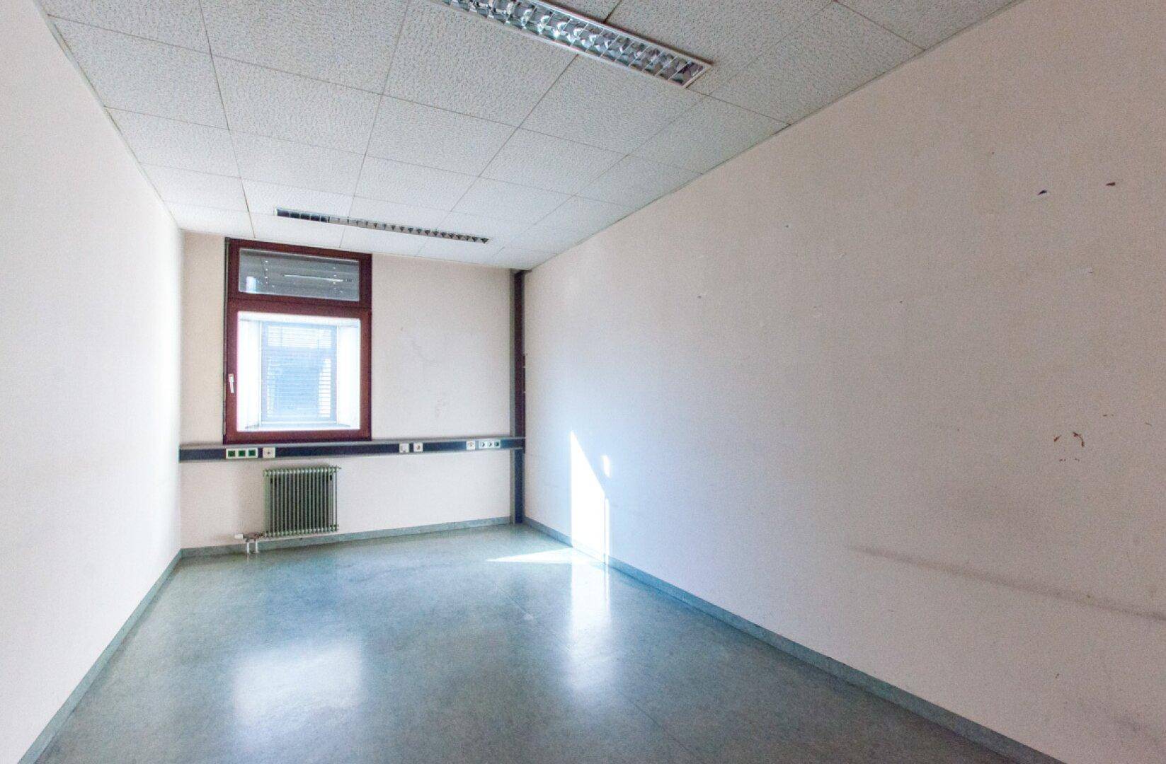 Schmaler Büroraum mit einem Fenster, weißen Wänden und einem glänzenden grauen Boden, der das Licht reflektiert.