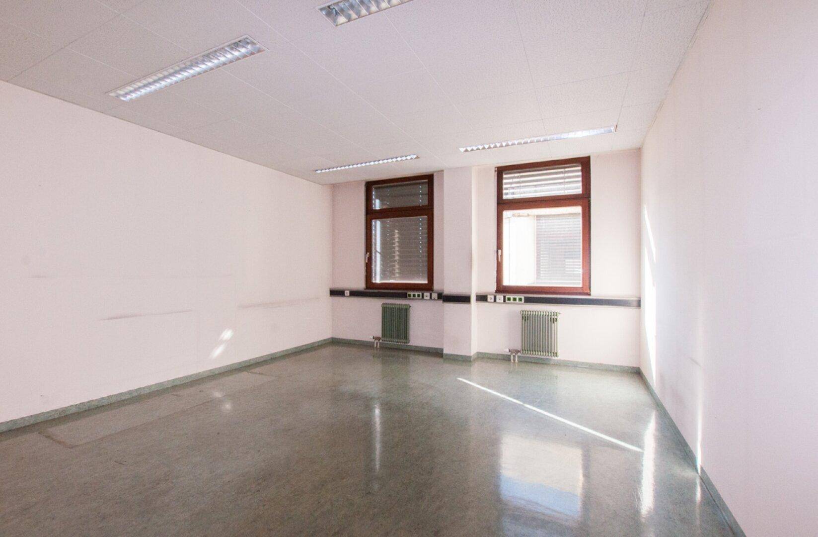 Helles Büro mit zwei Fenstern, weißen Wänden und einem glänzenden grauen Boden, der das Sonnenlicht reflektiert.