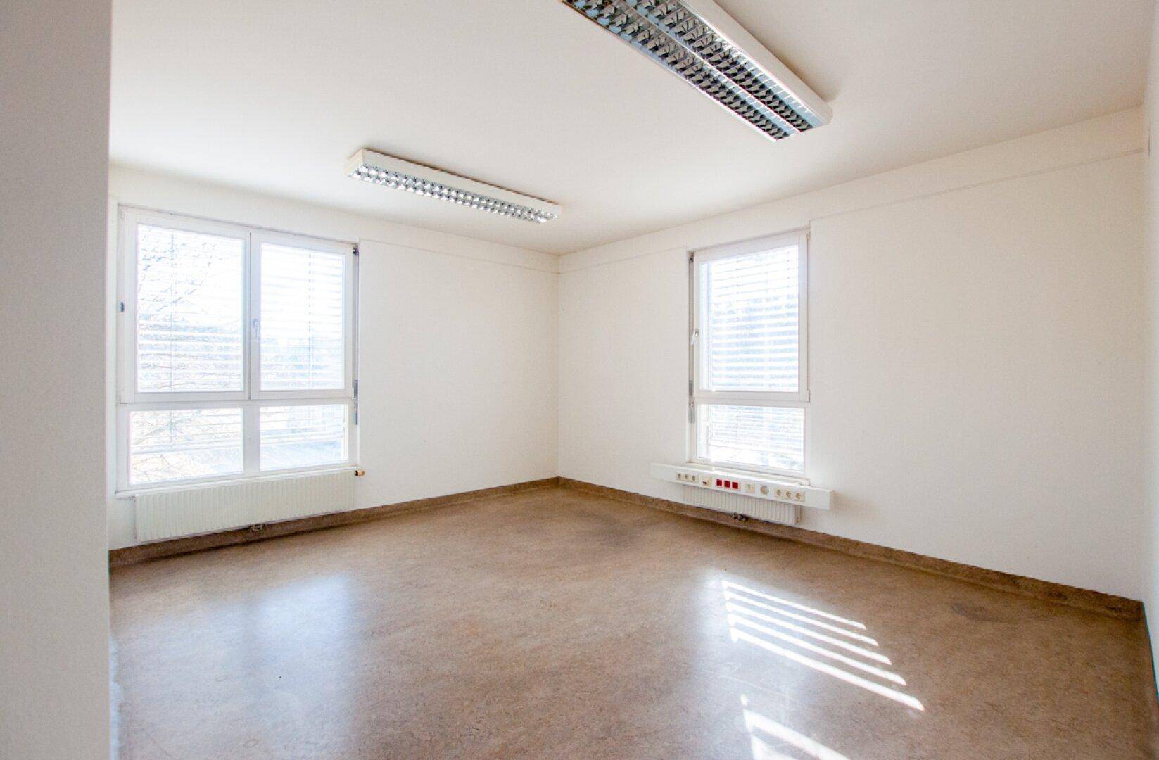 Geräumiges Büro mit zwei großen Fenstern, Linoleumboden und viel Tageslicht.