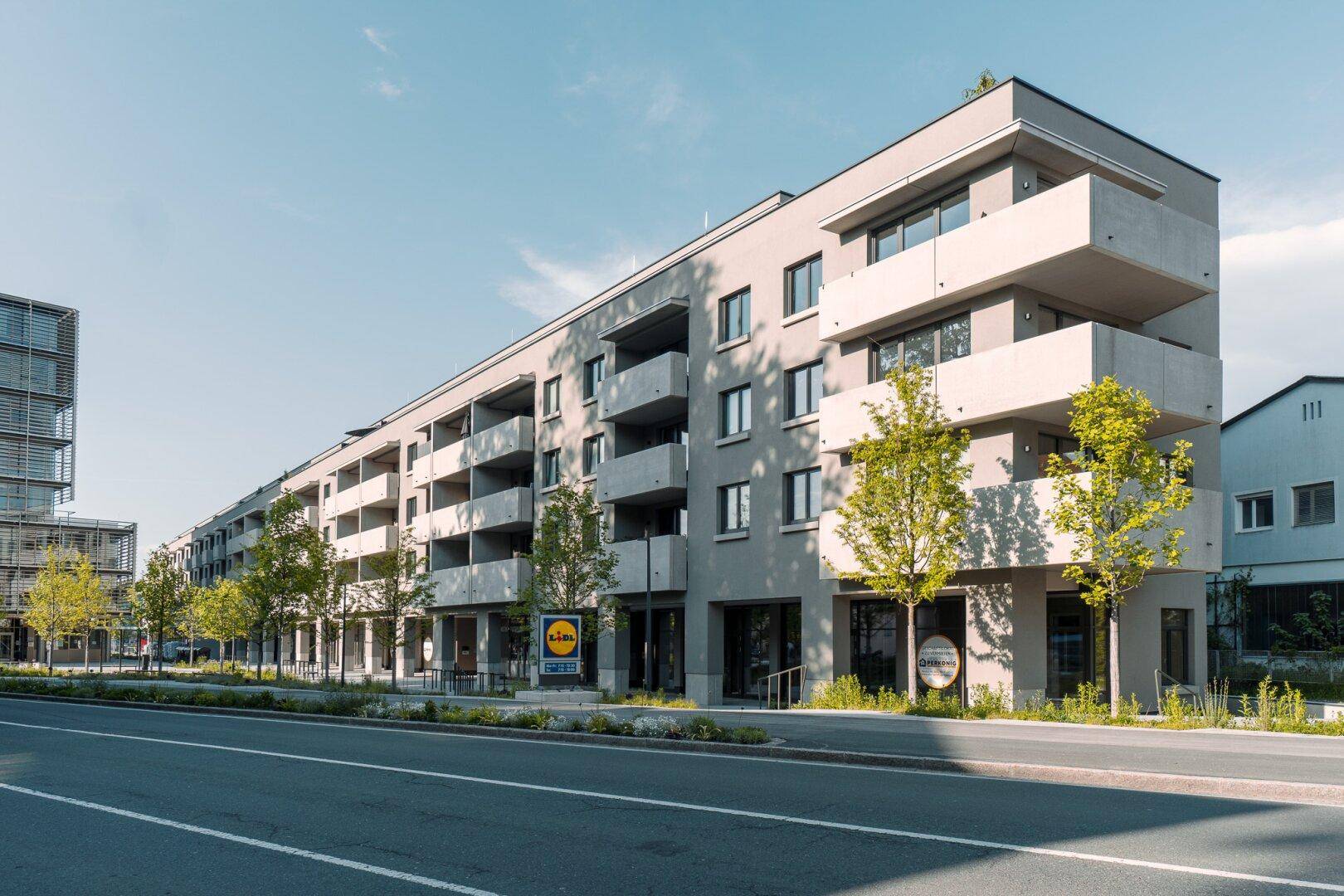 Lange Fassade eines modernen Wohngebäudes mit vielen Balkonen entlang einer Straße, umgeben von Bäumen.