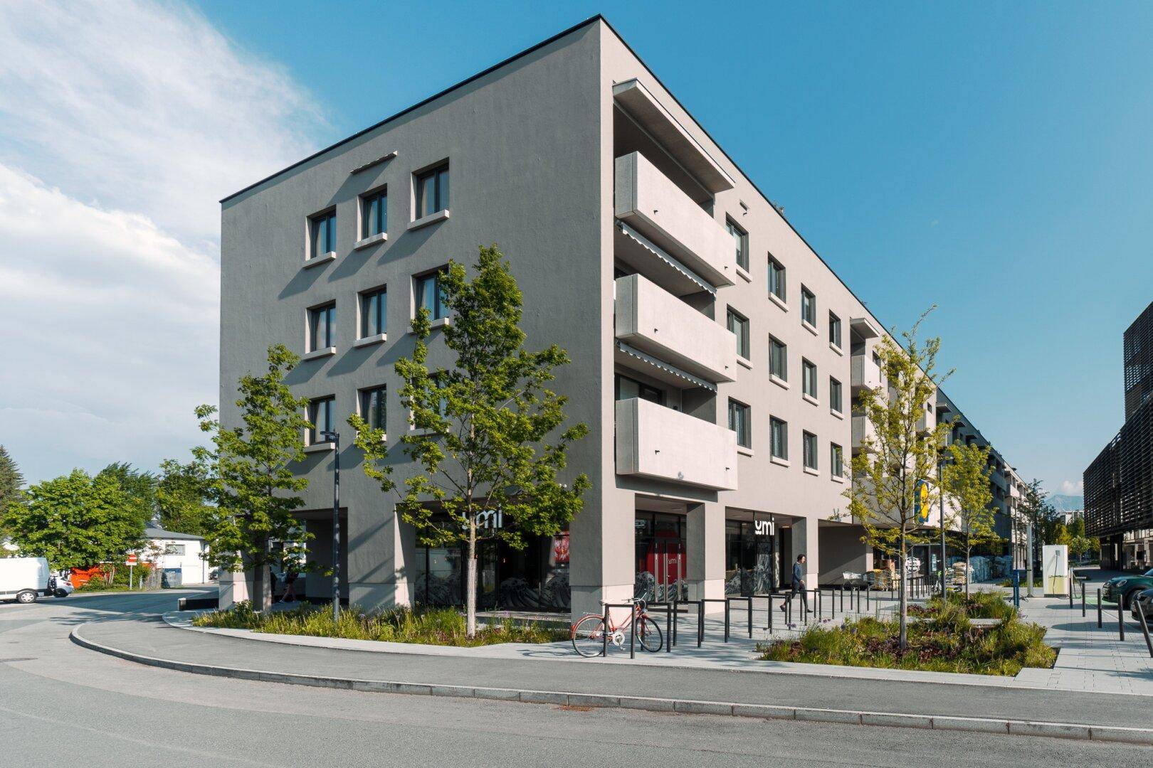Modernes Mehrfamilienhaus mit grauer Fassade, Balkonen und Geschäftsflächen im Erdgeschoss an der Straße.