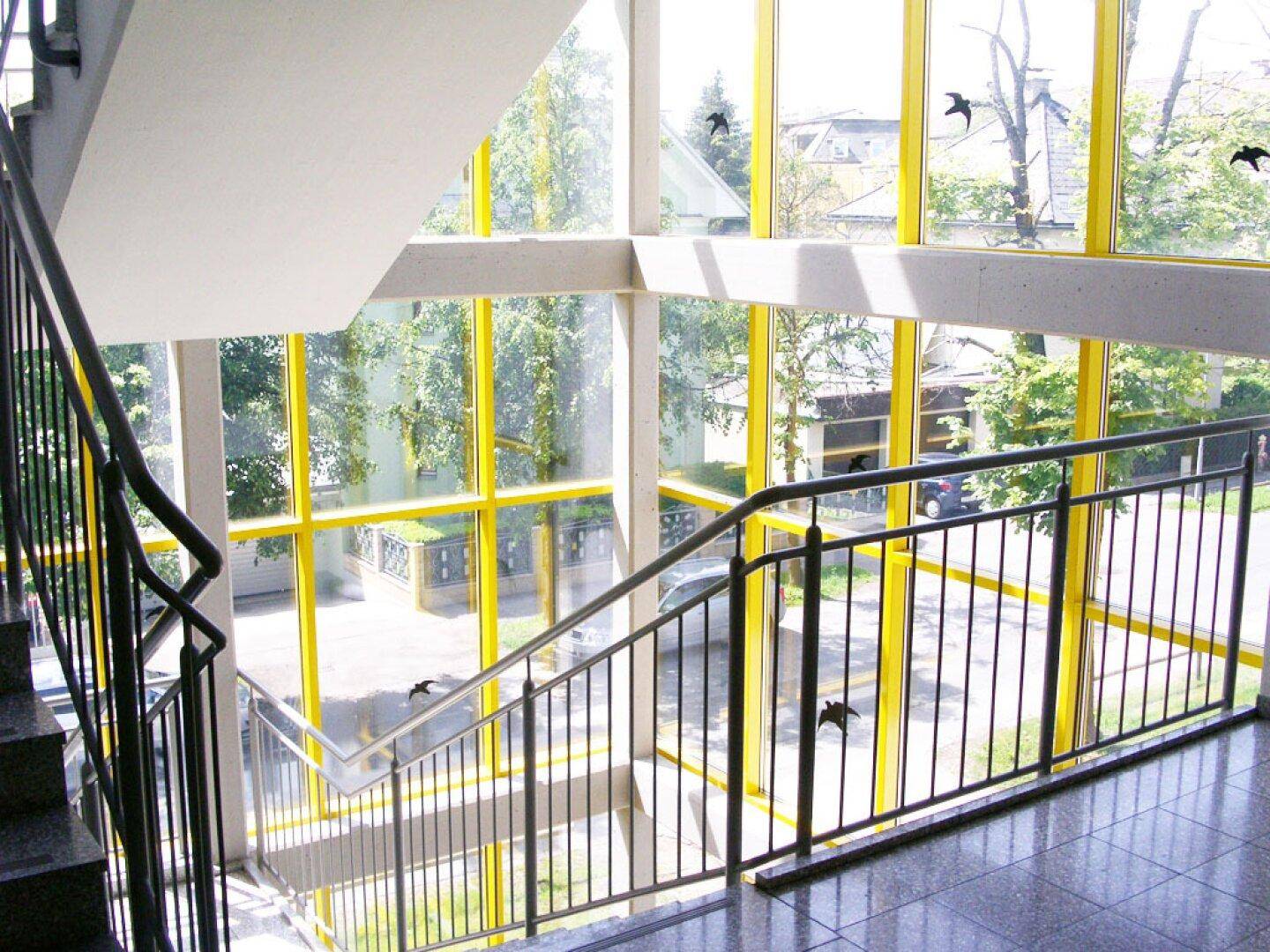 Moderner Treppenbereich mit großen gelben Fensterfronten, die viel Tageslicht hereinlassen und einen Blick ins Grüne bieten.