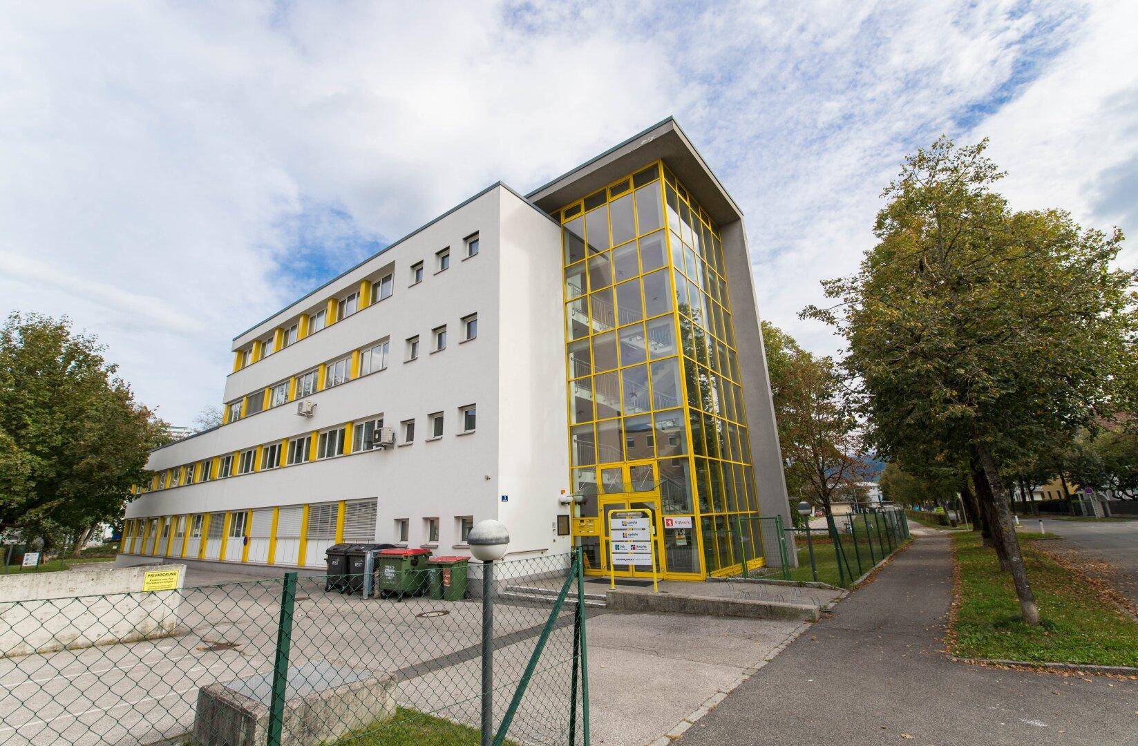 Ansicht des modernen Bürogebäudes mit gelben Fensterrahmen und großem Glas-Eingangsbereich unter bewölktem Himmel.