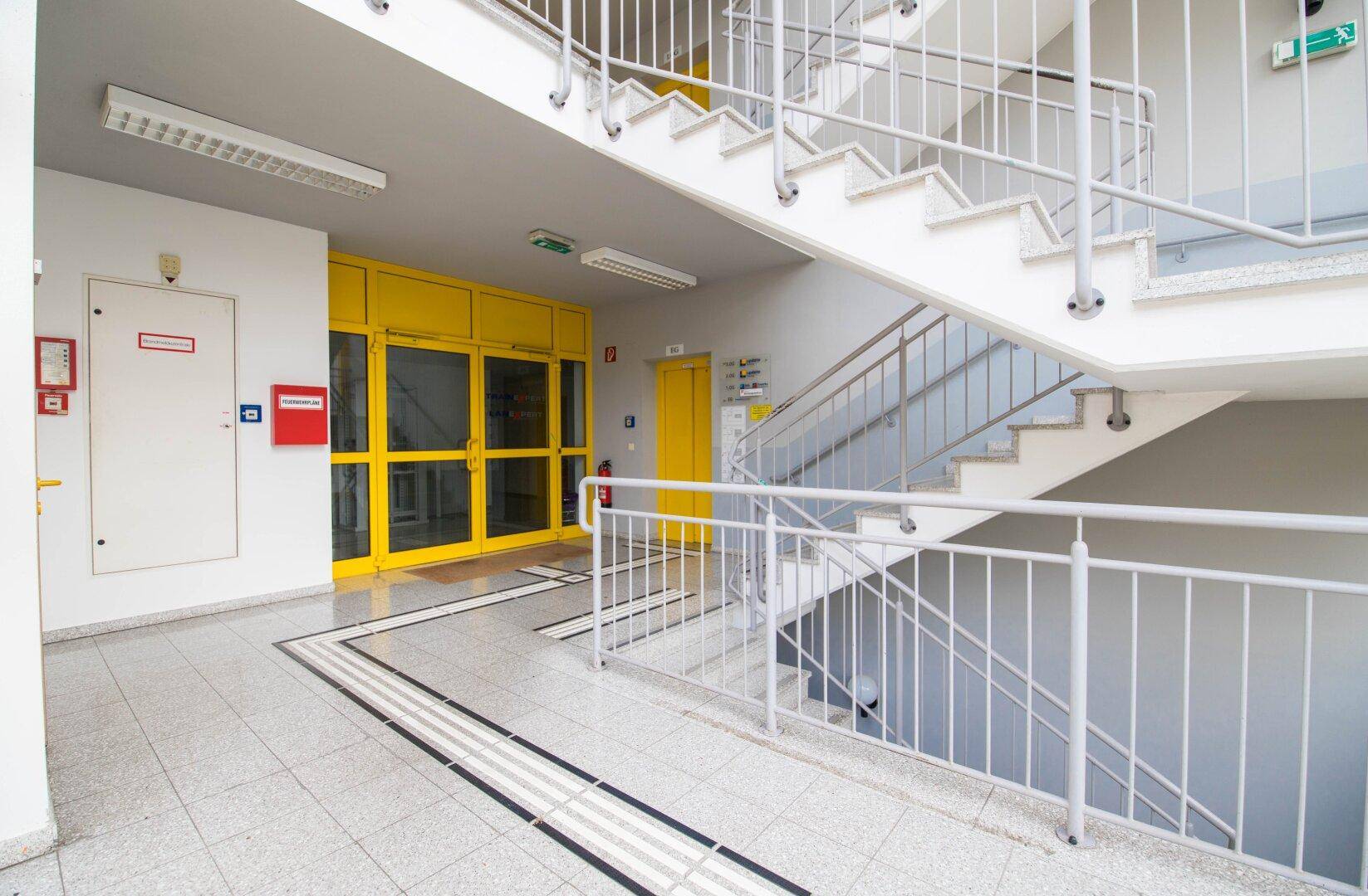 Heller Treppenaufgang mit Metallgeländer und Fliesenboden, angrenzend an einen Eingangsbereich mit gelben Türen.