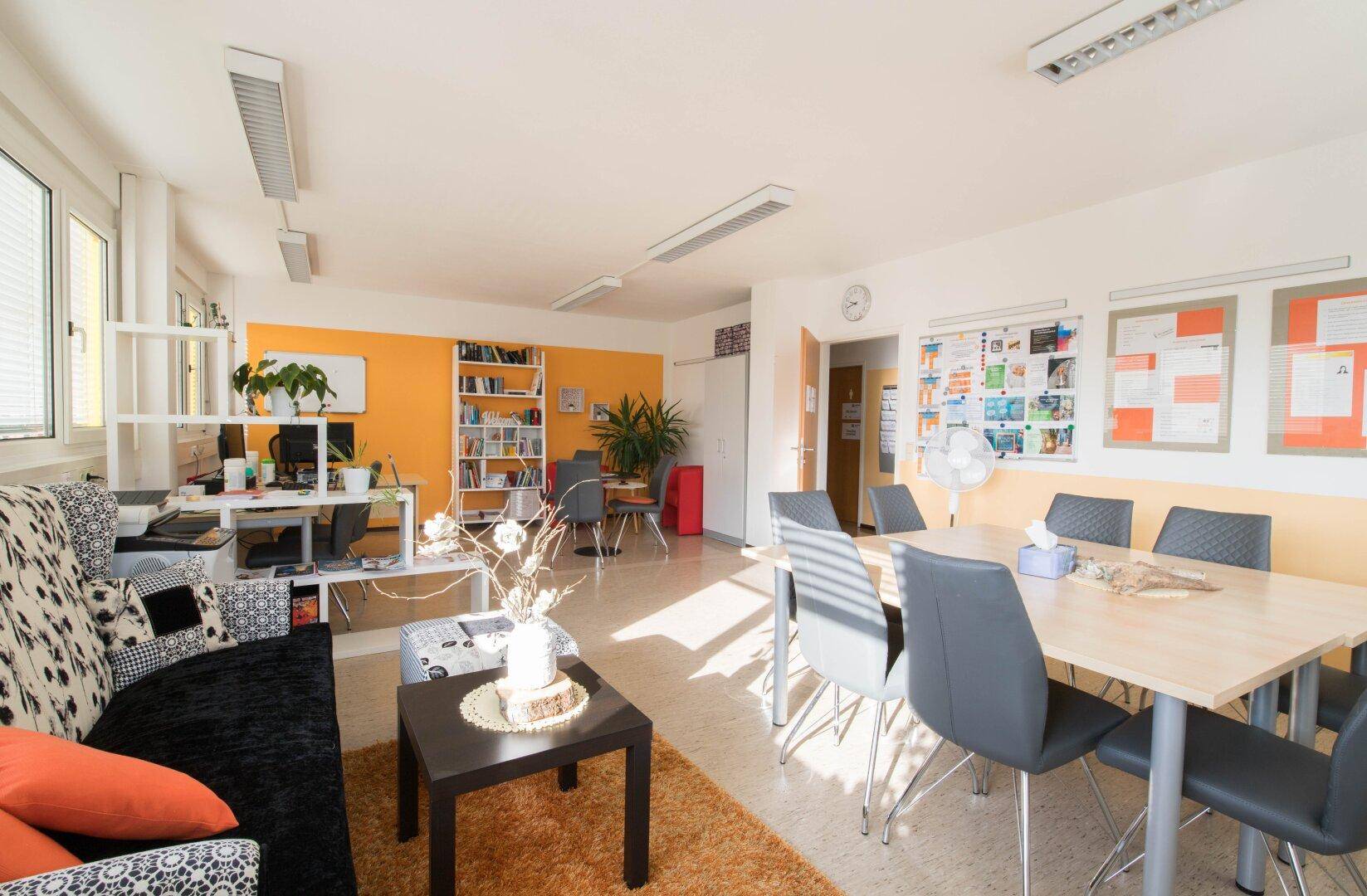 Geräumiger Gemeinschaftsraum mit Linoleumboden, orangefarbener Akzentwand und verschiedenen Arbeits- und Sitzbereichen.