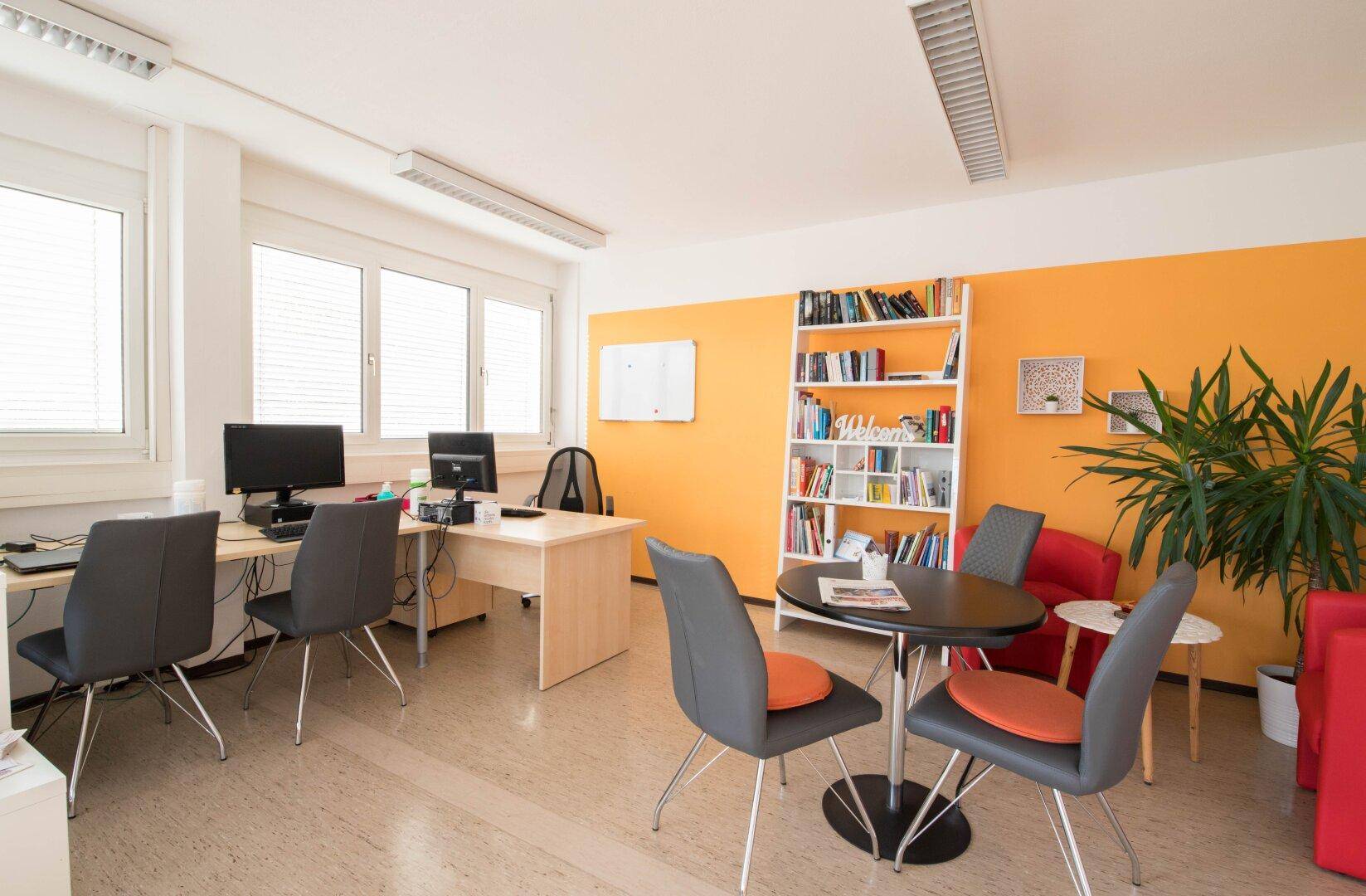 Helles Großraumbüro mit Linoleumboden, orangefarbener Wand und mehreren Arbeitsplätzen sowie einem Bücherregal.