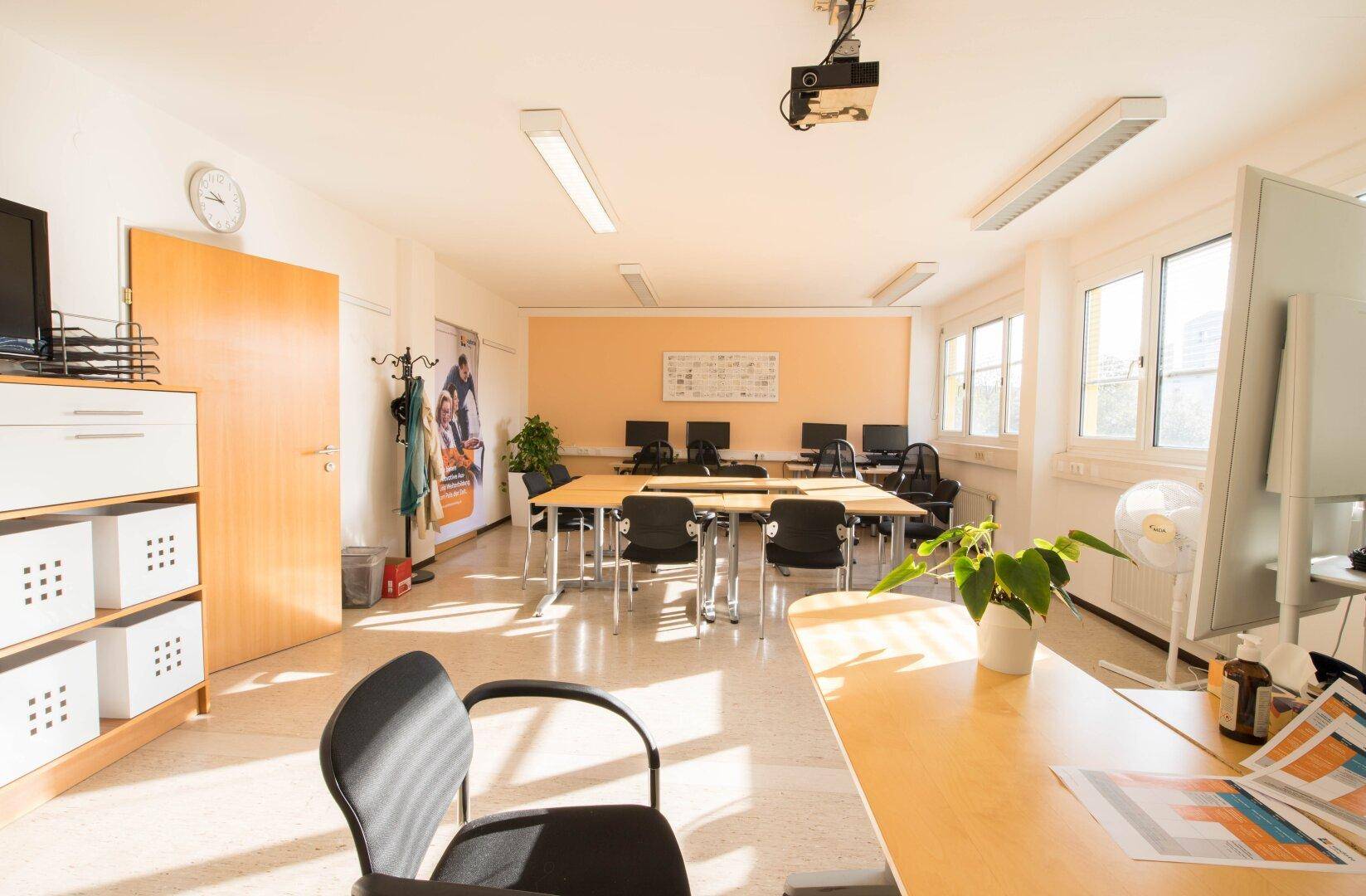 Heller Schulungsraum mit Linoleumboden, vielen Fenstern und mehreren Tischen und Stühlen für Unterrichtszwecke.