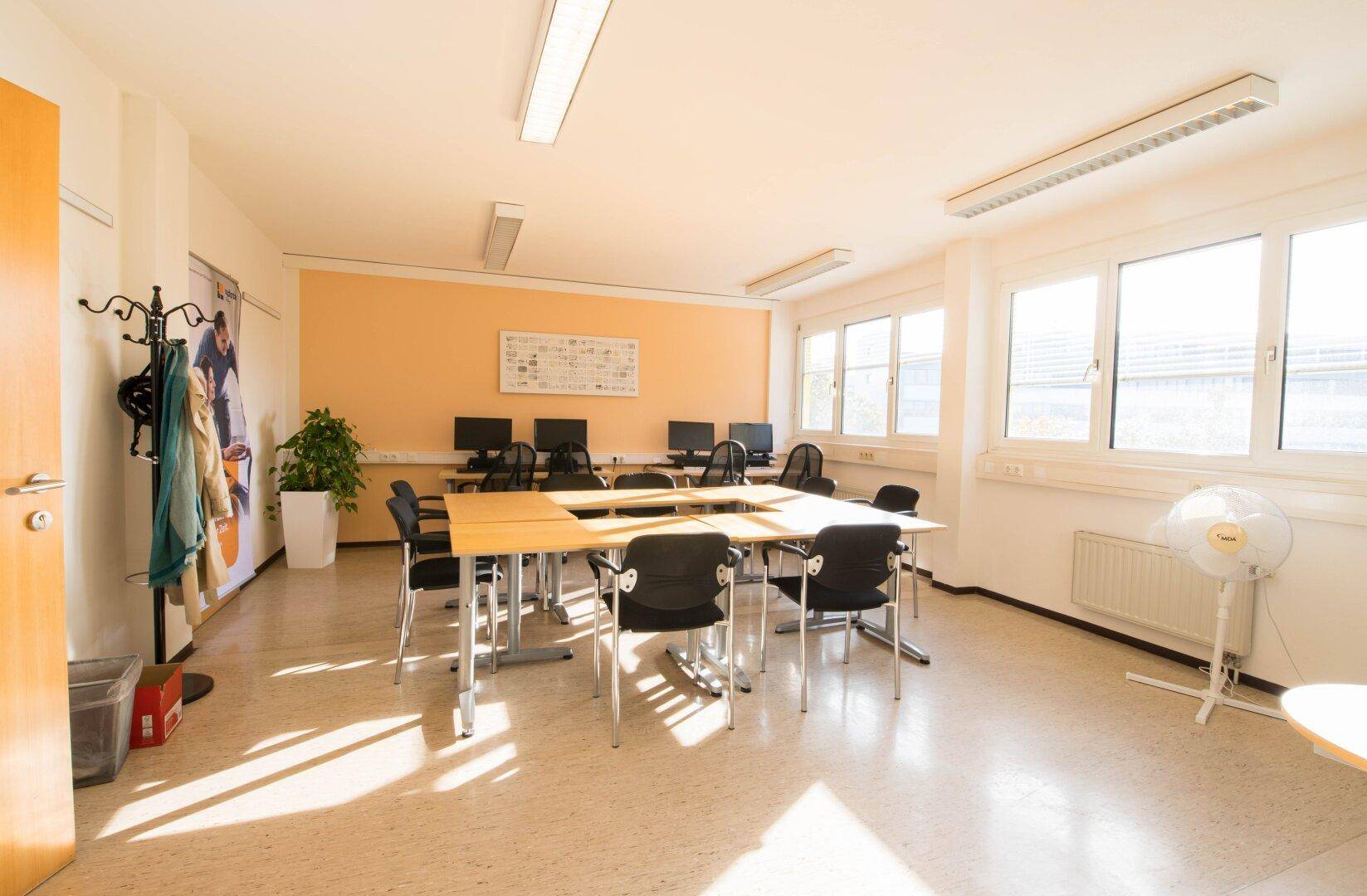 Geräumiger Schulungsraum mit Linoleumboden, großen Fenstern und mehreren Tischen, ideal für Gruppenarbeiten.