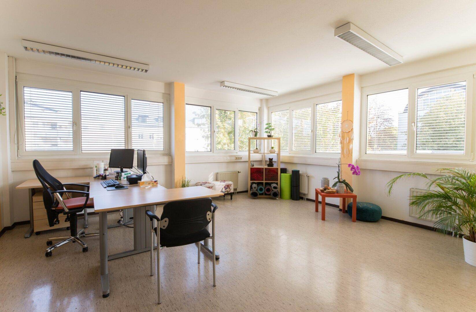 Vielseitiges Büro mit Linoleumboden, großen Fenstern und einem Arbeitsplatz, auch für Yoga-Übungen nutzbar.