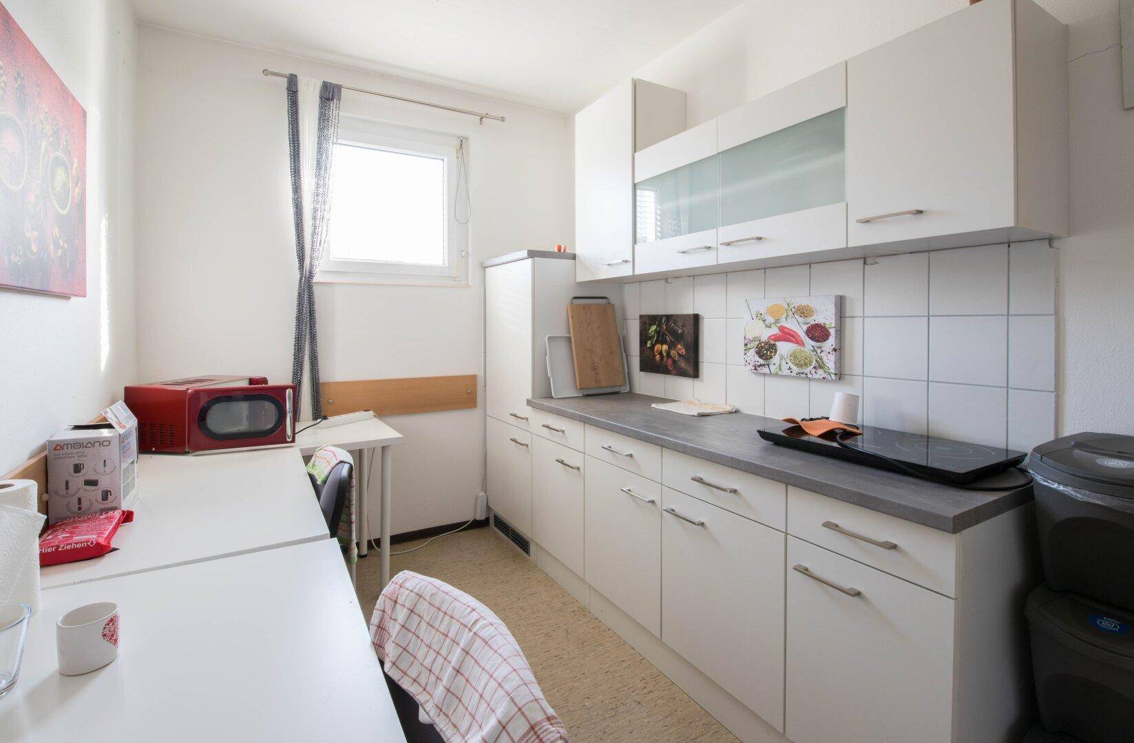 Funktionale Küche mit weißen Schränken, grauer Arbeitsplatte und einem Fenster, ausgestattet mit Mikrowelle und Kochfeld.