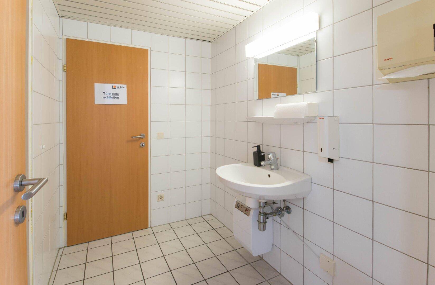 Einfache Toilette mit weißen Fliesen, Waschbecken und Holztür, ausgestattet mit Seifenspender und Spiegel.