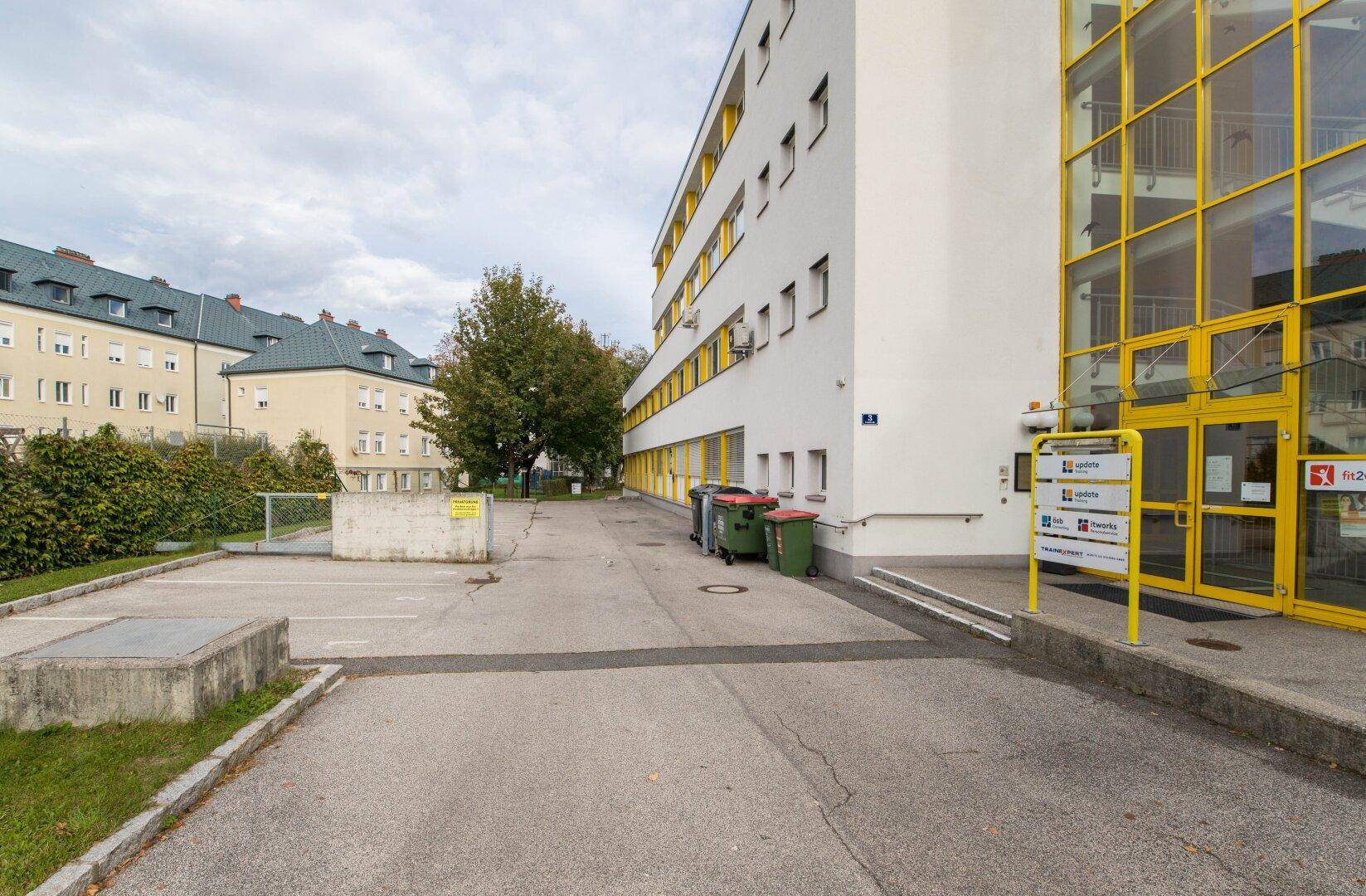 Außenansicht eines modernen Gebäudes mit gelben Akzenten, Parkplätzen und Mülltonnen im Vordergrund.