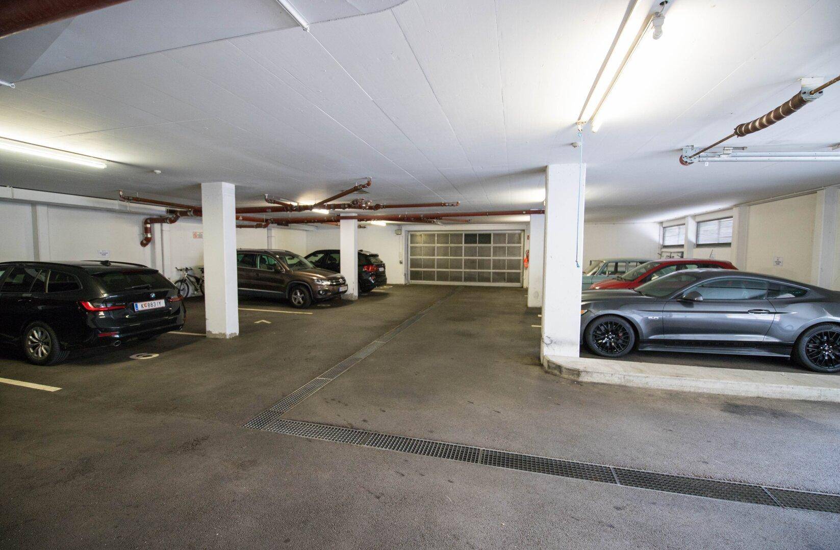 Geräumige Tiefgarage mit mehreren geparkten Autos und sichtbaren Versorgungsleitungen an der Decke.