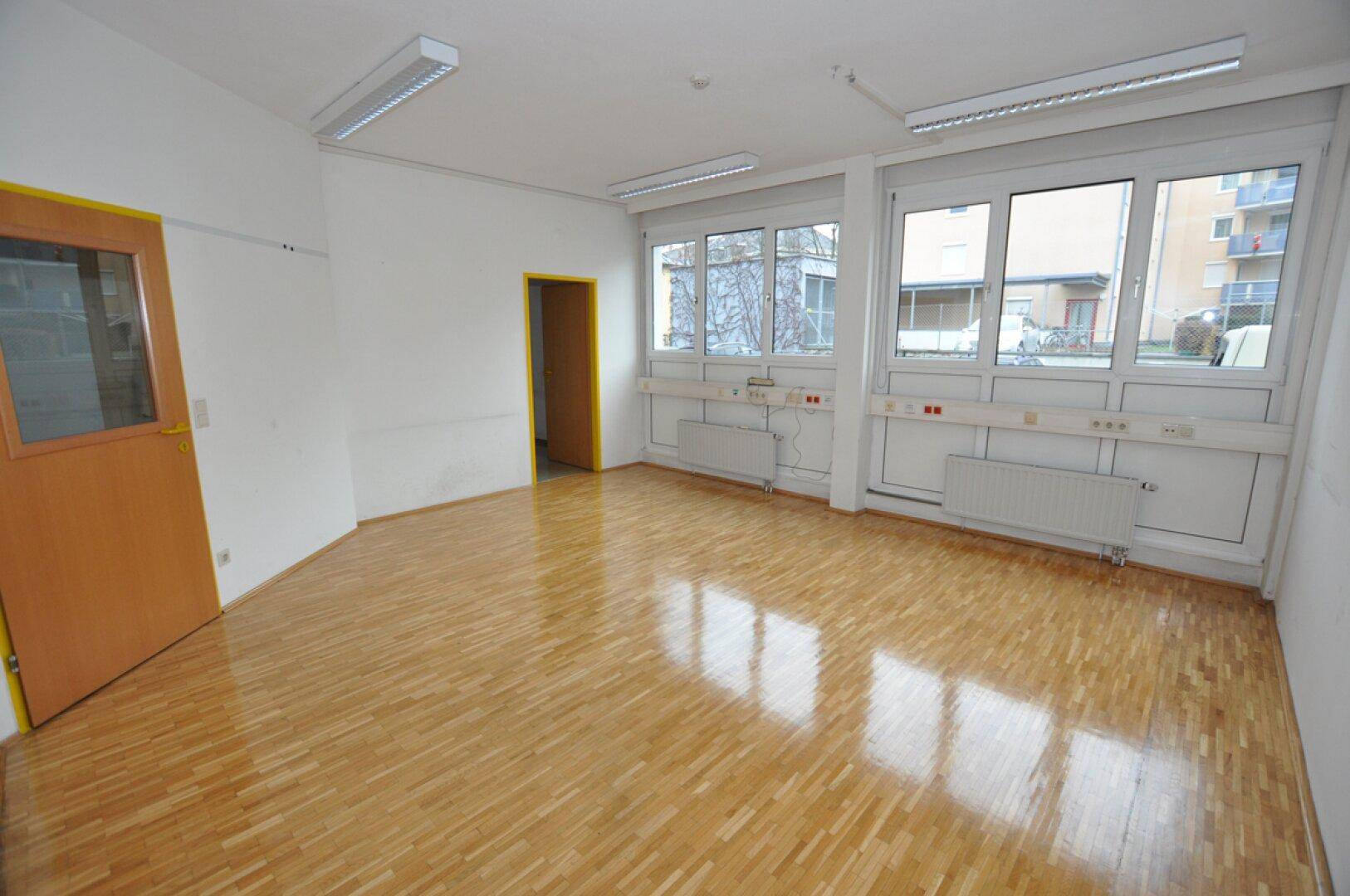 Leeres Büro mit großen Fenstern, Holzparkettboden und Heizkörpern unter den Fenstern.