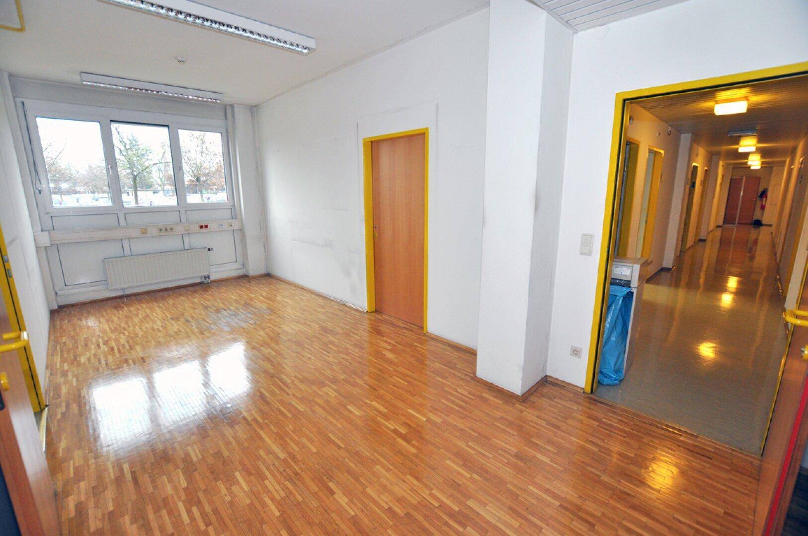 Geräumiger Büroraum mit glänzendem Holzparkettboden und einem Eingang zu einem langen Flur.
