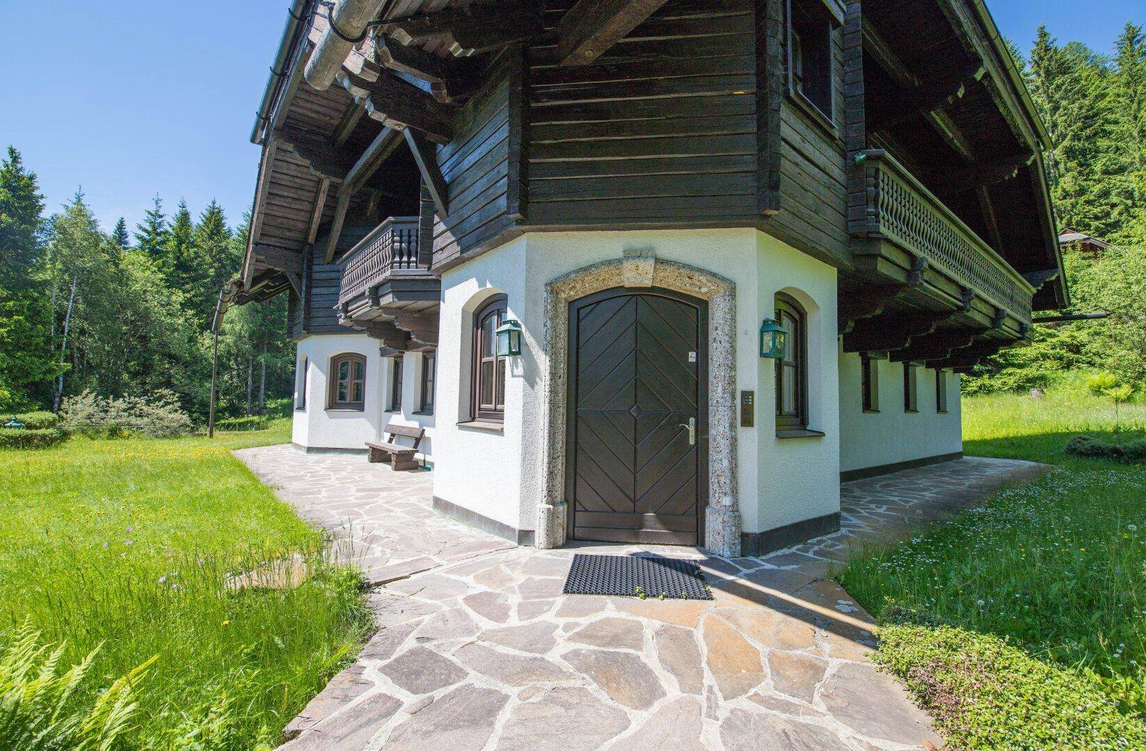 Rustikaler Haupteingang des Chalets mit Holztür und Natursteinumrandung, umgeben von gepflegten Grünflächen.