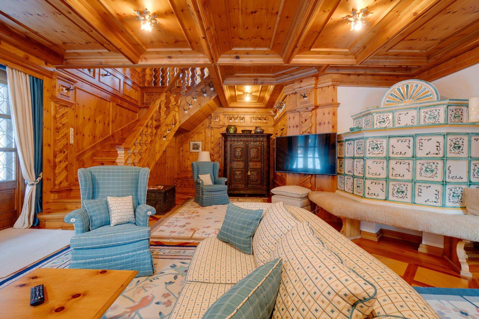 Gemütlicher Wohnbereich mit großem Fernseher, traditionellem Kachelofen und Holztreppe, die den Raum verbindet.