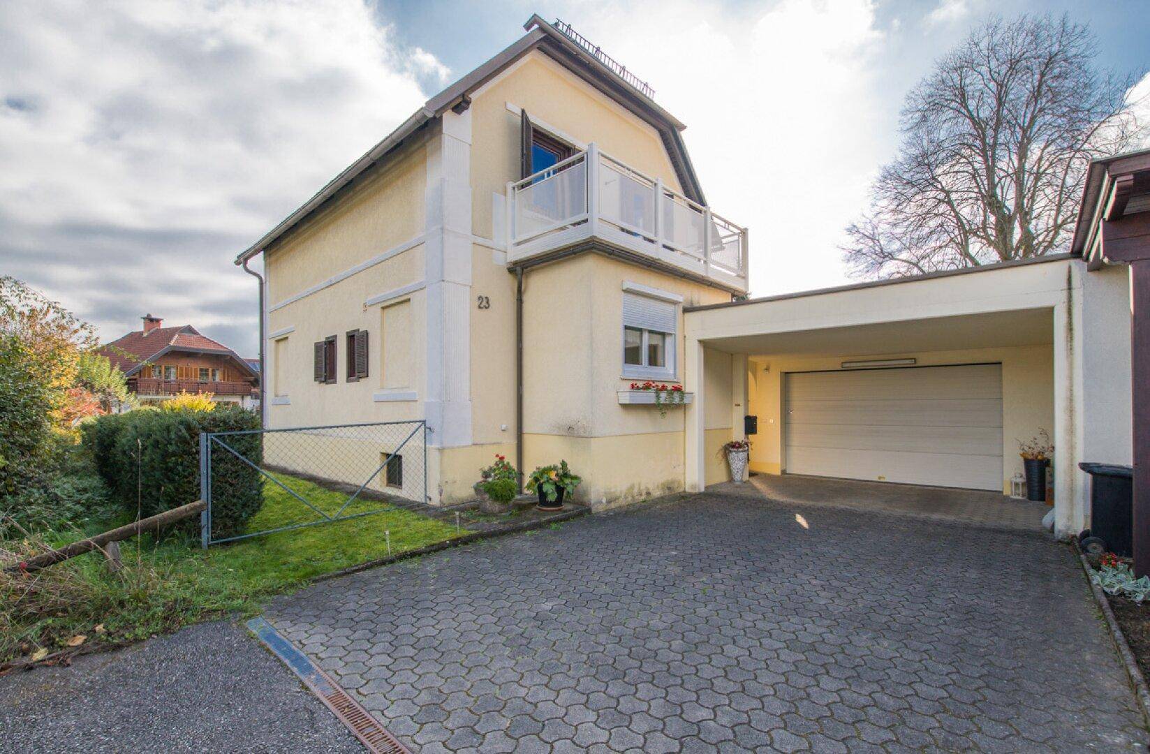 Vorderansicht des Hauses mit gepflasterter Auffahrt, Carport und Balkon im Obergeschoss.