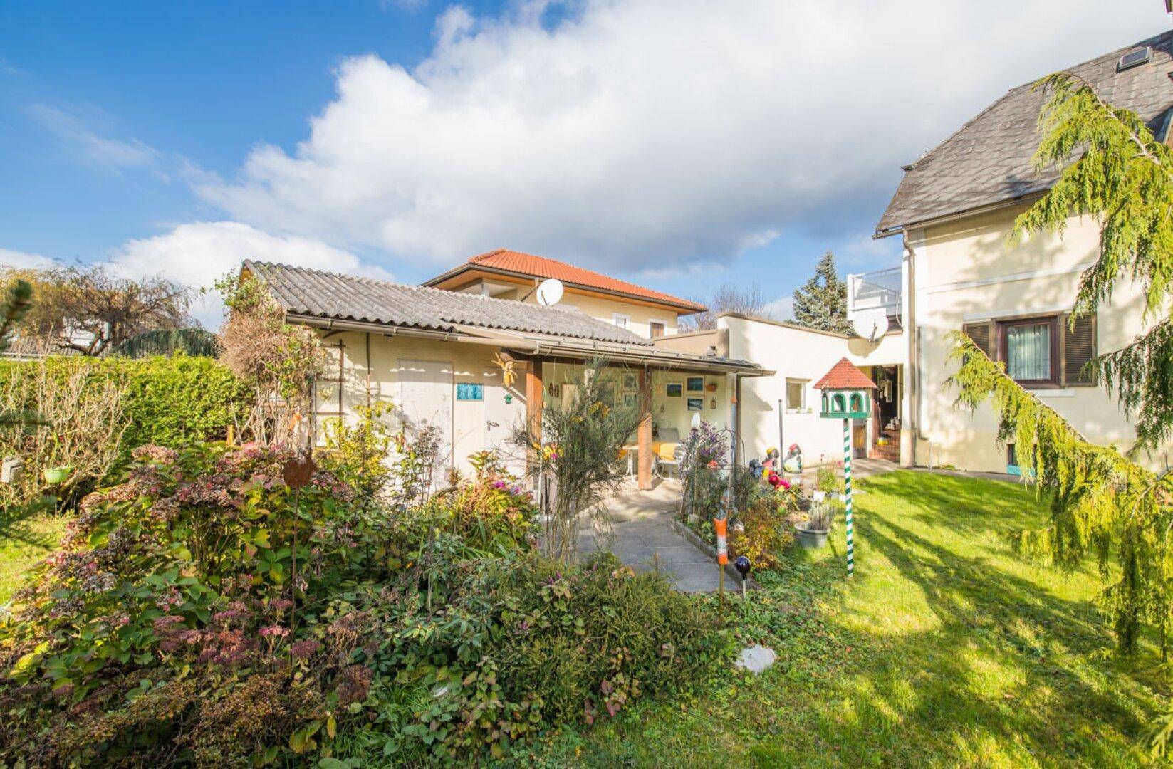 Blick auf das Haus und ein Nebengebäude mit überdachtem Sitzbereich im grünen Garten.