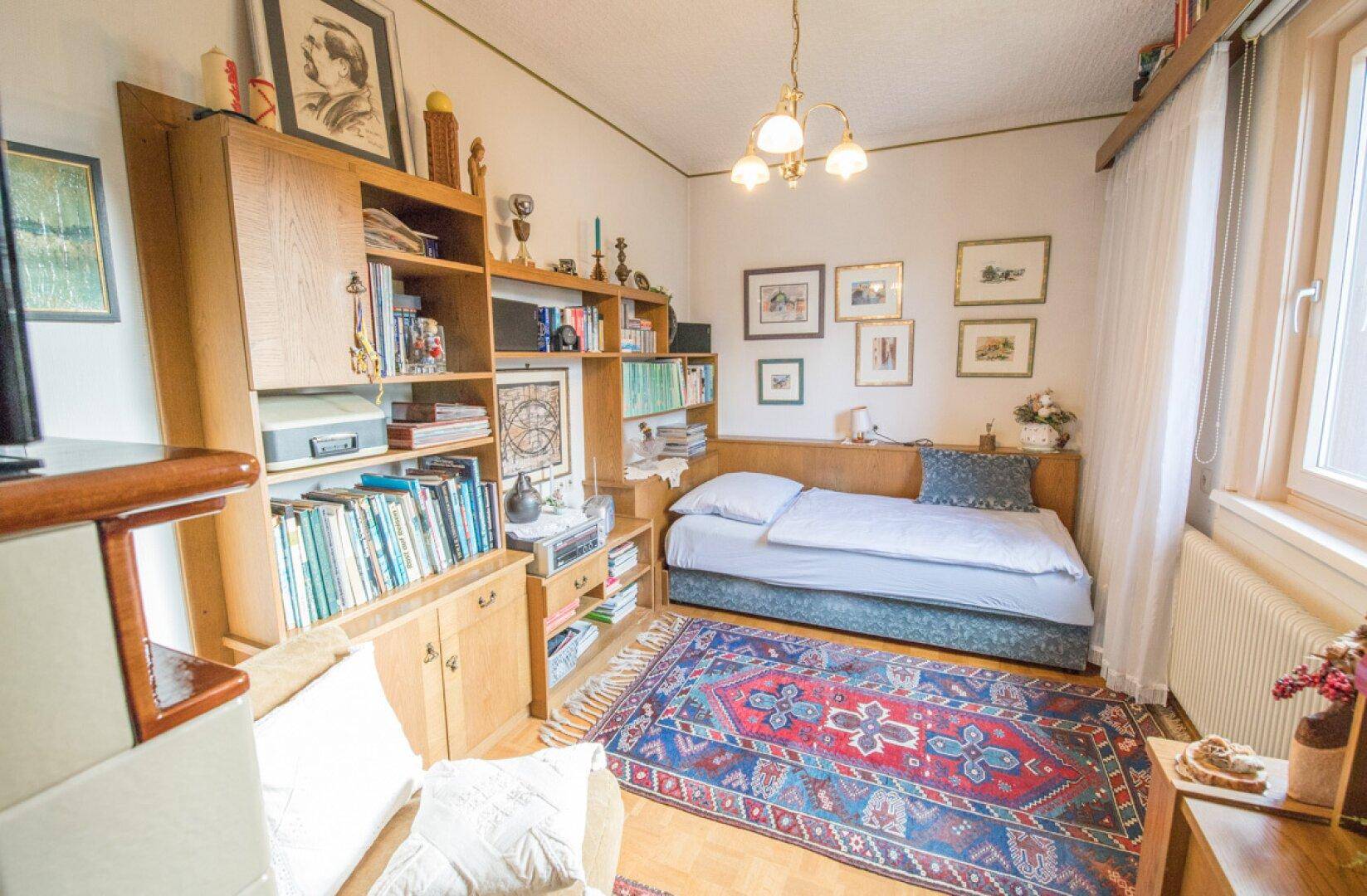 Schlafzimmer mit Bücherregal, Kachelofen und traditioneller Einrichtung.