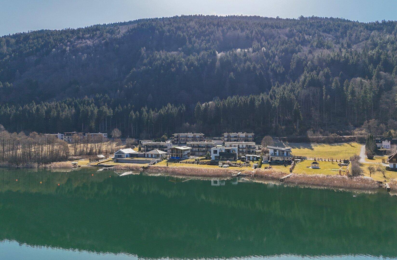 Luftaufnahme einer Wohnanlage am Seeufer mit bewaldeten Bergen im Hintergrund.