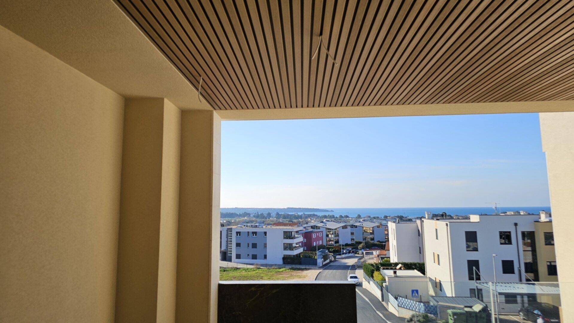 Blick von einem Balkon mit Holzlamellendecke auf das Meer und die umliegende Bebauung.