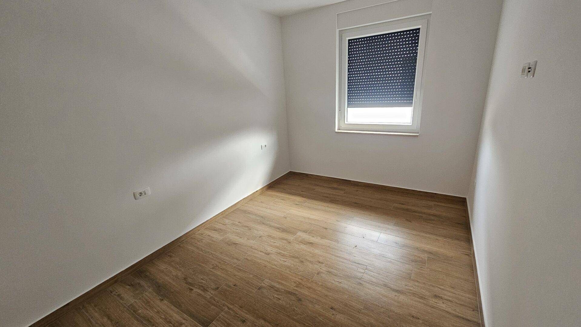 Helles, leeres Zimmer mit Laminatboden und Fenster mit Rollladen.