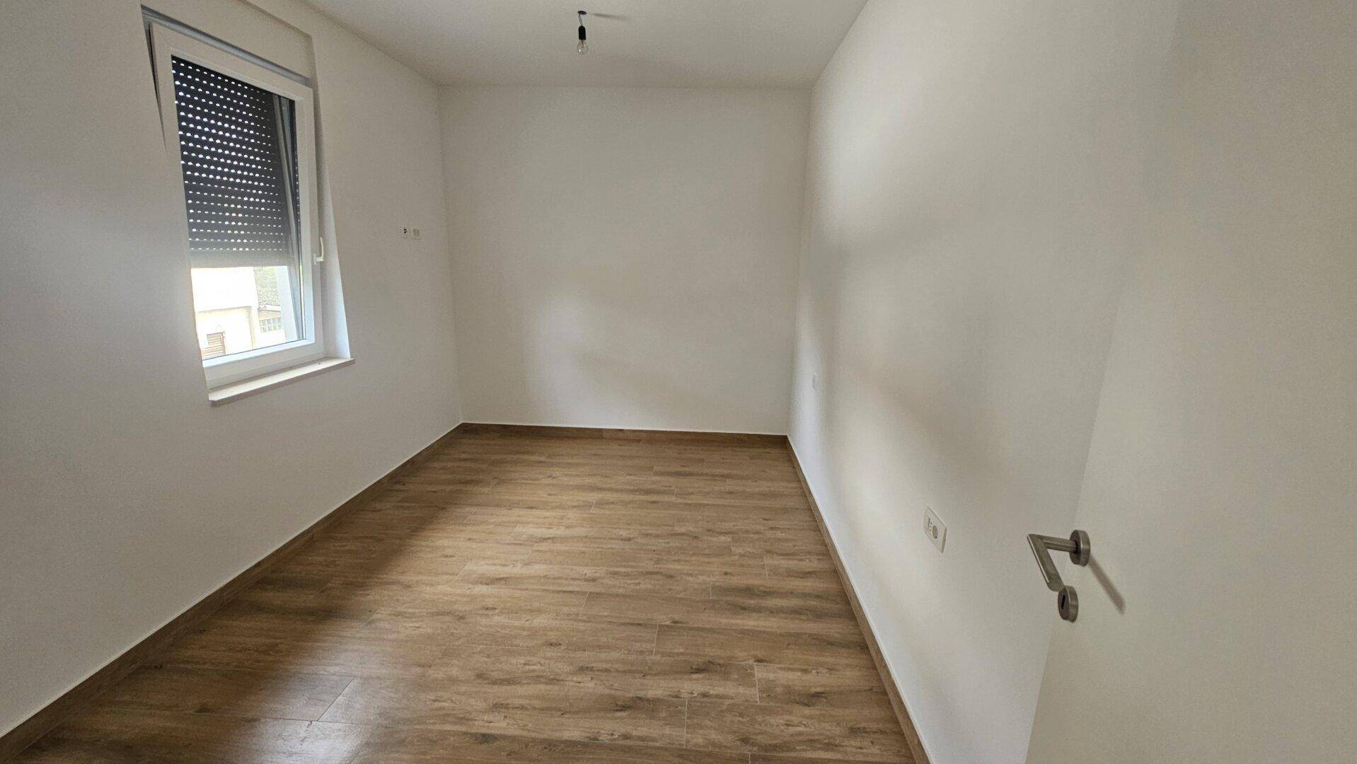 Leeres Zimmer mit Laminatboden und Fenster mit geschlossenem Rollladen.