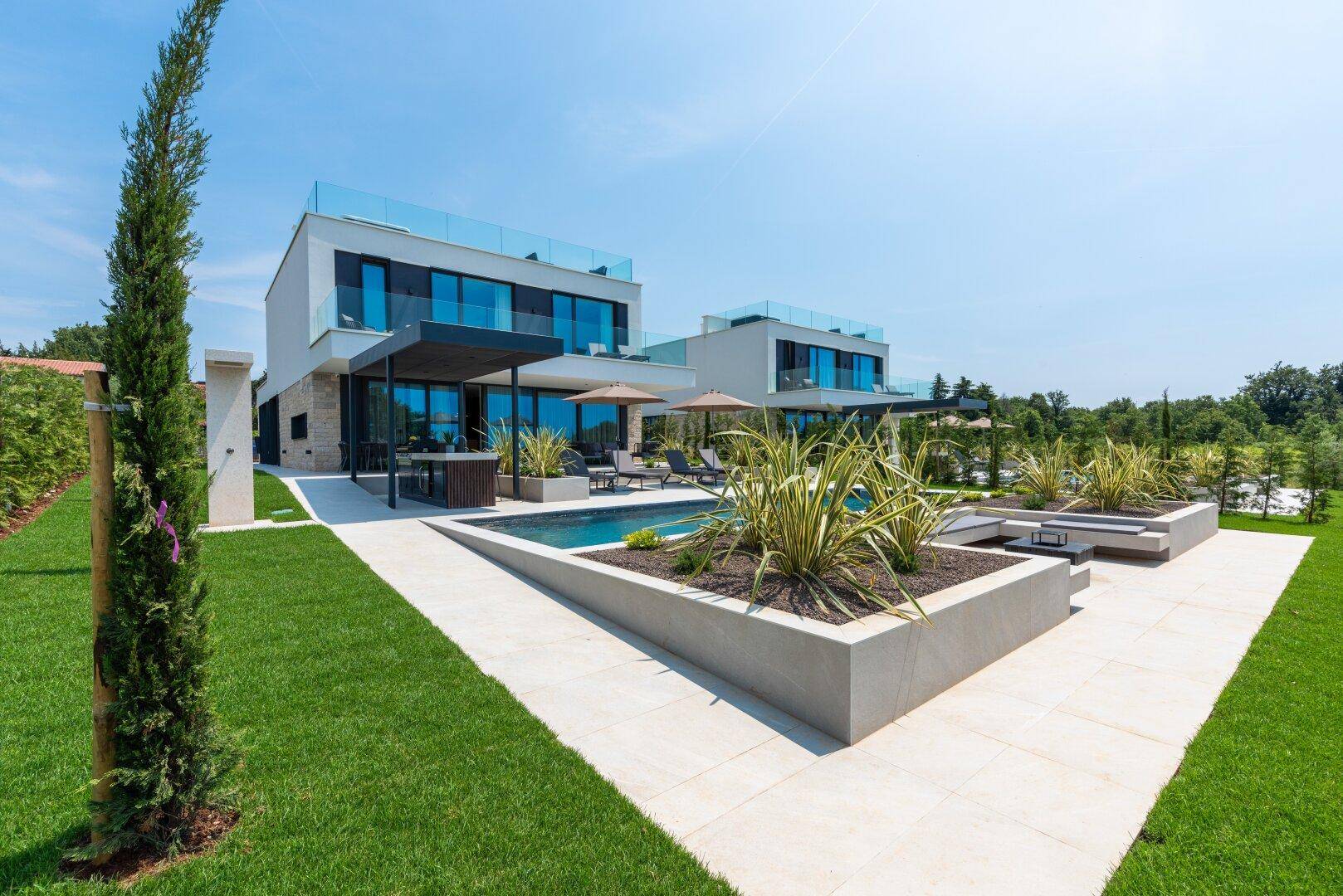 Moderne Villa mit Poolbereich, Sonnenterrasse und Blick ins Grüne unter blauem Himmel.